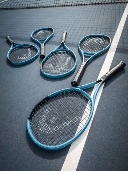 Five blue tennis rackets, including a HEAD Boom Team 2026 Tennis Racket, lie casually on the blue hard court near the white boundary line and net, with their handles and strings overlapping.