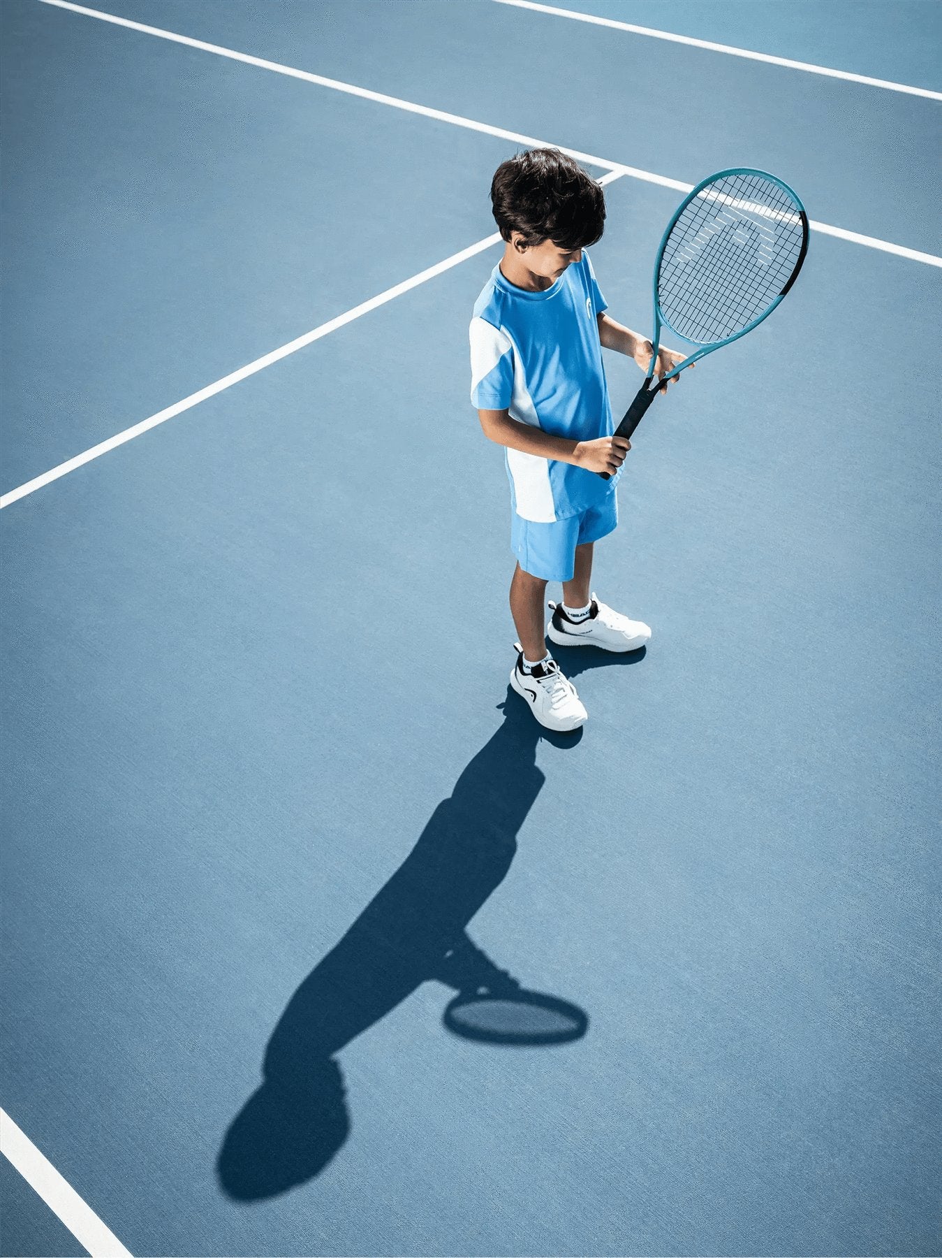 A young boy in a blue athletic outfit stands on a blue tennis court, holding a HEAD Boom Jr 2026 Junior Tennis Racket by HEAD, looking down as his shadow is cast clearly on the court beside him.