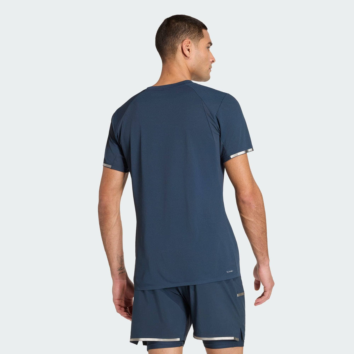 A man wearing the adidas Climacool+ Freelift Pro Mens Tennis T-Shirt - Aurora Ink stands with his back to the camera against a plain light gray background.