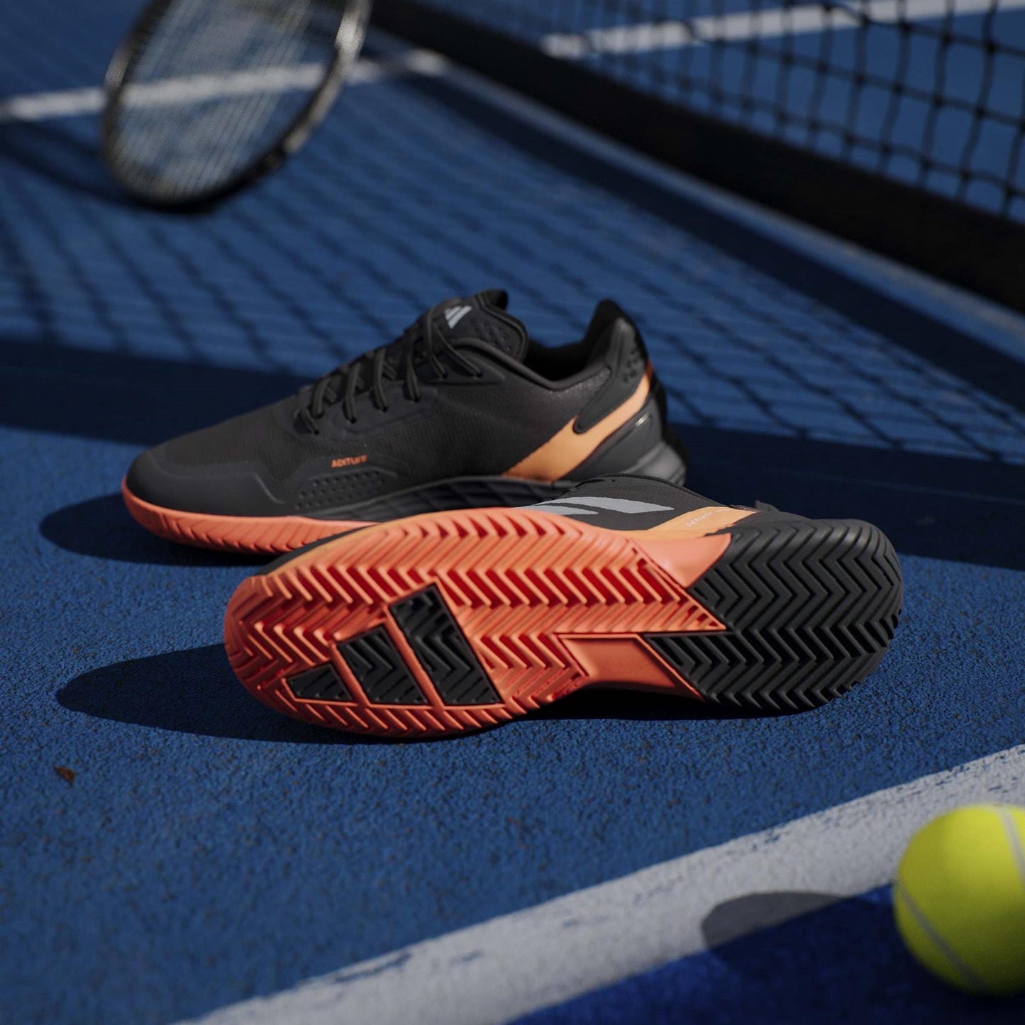 A pair of adidas Defiant Speed 2 Men's Tennis Shoes in Black/Orange is displayed on a blue tennis court, with a racket and tennis ball nearby.