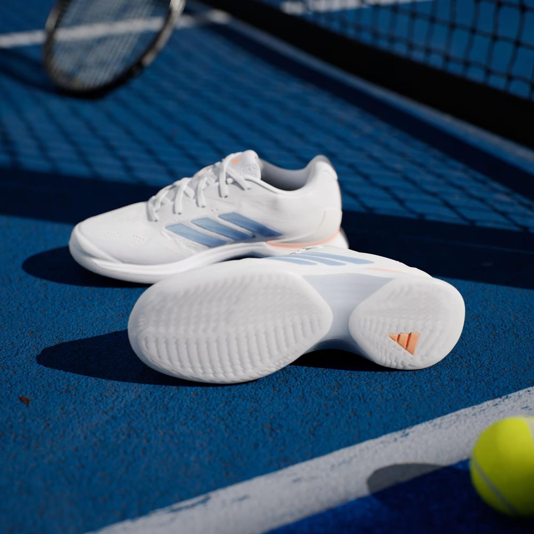 A pair of adidas ADIDAS Avacourt 3 Women’s Tennis Shoes in Core White, Halo Blue, and Beam Orange are on a blue tennis court next to a racquet and yellow tennis ball.