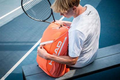 A tennis player in a white shirt sits on a blue court bench, unzipping the orange HEAD Tour 25L Tennis Backpack and holding a racquet while looking down.