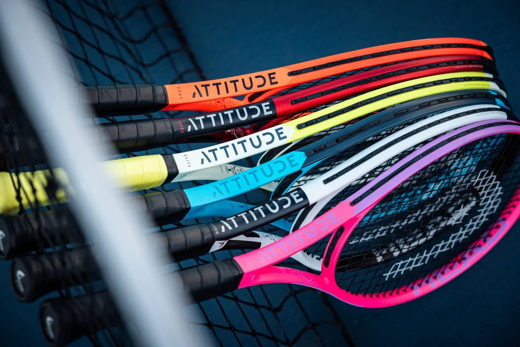 A row of HEAD MX Attitude SUPRM Tennis Rackets from HEAD, each in a unique vibrant color with "ATTITUDE" on the frame, rests against a tennis net—perfect beginner rackets for standout style on the court.