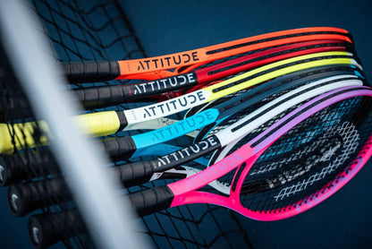 A row of HEAD MX Attitude SUPRM Tennis Rackets from HEAD, each in a unique vibrant color with "ATTITUDE" on the frame, rests against a tennis net—perfect beginner rackets for standout style on the court.