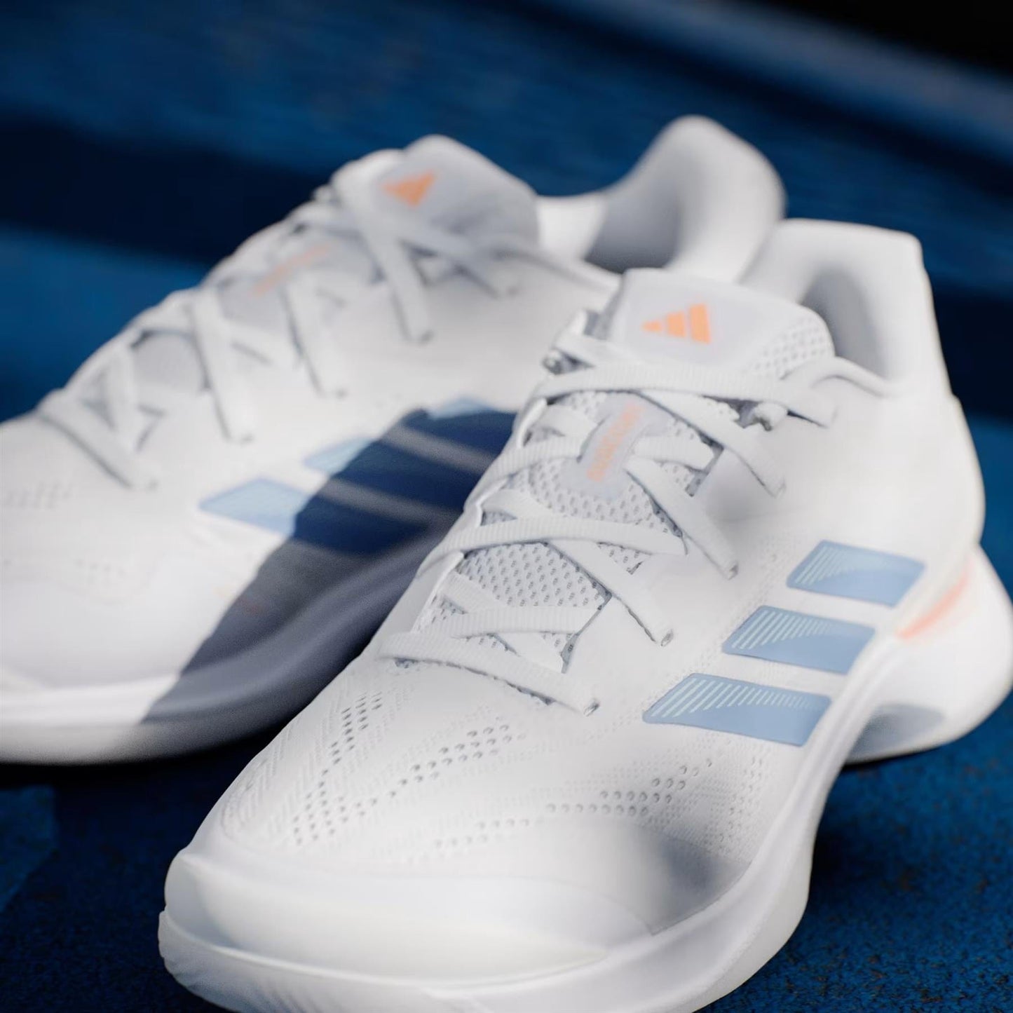 A close-up of the adidas Avacourt 3 women's tennis shoes in Core White, Halo Blue, and Beam Orange shows detailed laces and side stripes on a blue surface.