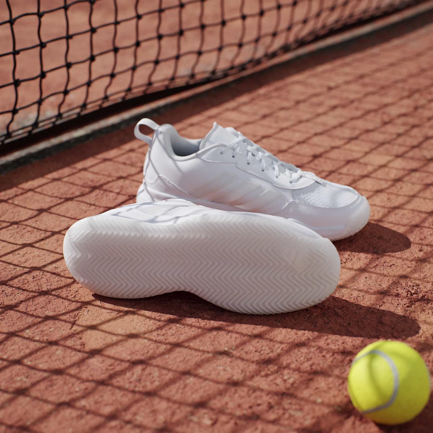 A pair of adidas AvaFlash Women's Tennis Shoes - White rests on a clay court near a tennis ball, as the net casts delicate shadows over the lightweight shoes.