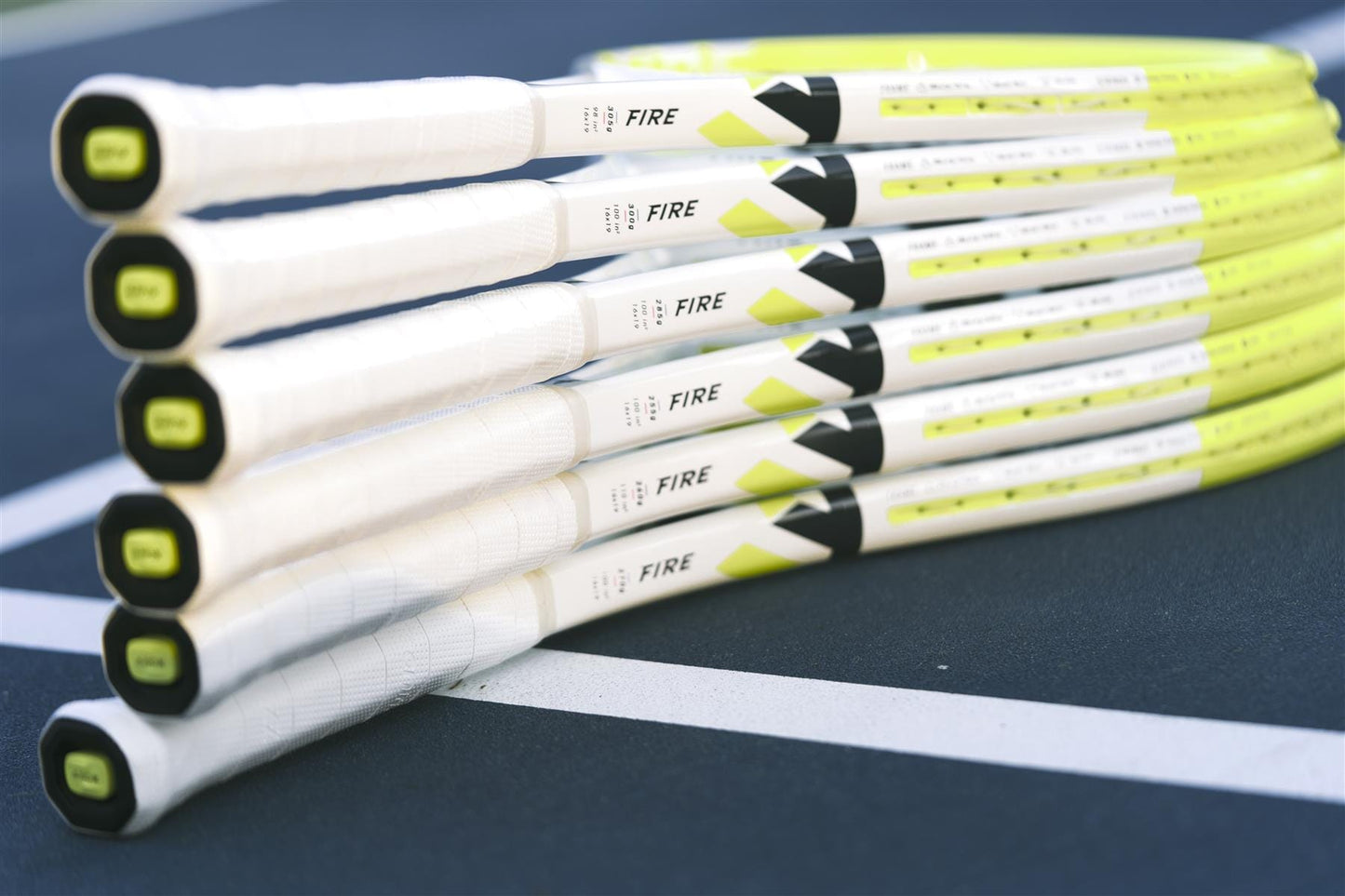 A close-up of seven Tecnifibre Fire 255 Tennis Rackets stacked with handles facing forward on a blue court, showcasing their yellow and white design and advanced X-Damp technology for superior vibration dampening.