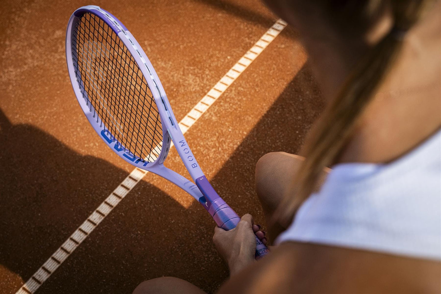 A close-up from behind shows a tennis player gripping the HEAD Boom MP Alternate 2026 Tennis Racket on a clay court, with their white outfit and arm visible. White court lines highlight the power of HEAD’s latest tennis racket in action.