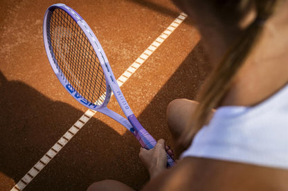 A close-up from behind shows a tennis player gripping the HEAD Boom MP Alternate 2026 Tennis Racket on a clay court, with their white outfit and arm visible. White court lines highlight the power of HEAD’s latest tennis racket in action.