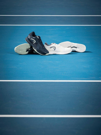 Two pairs of HEAD Sprint Team 4.0 Mens Tennis Shoes in Navy and Cantaloupe Green are placed side by side on a blue tennis court with visible white boundary lines.