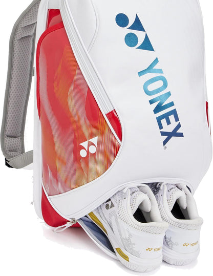 The Yonex 02612EX Expert Tennis Backpack in white features a fiery red and orange design with Yonex branding, plus a zippered bottom compartment where white Yonex sports shoes are partially visible.