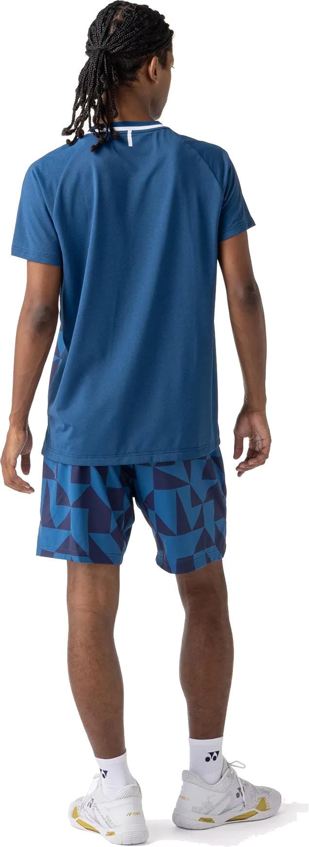 A person with braided hair stands facing away, wearing a blue t-shirt, Yonex 15240YX Knit Mens Tennis Shorts - Indigo Blue, white socks, and white athletic shoes against a white background.