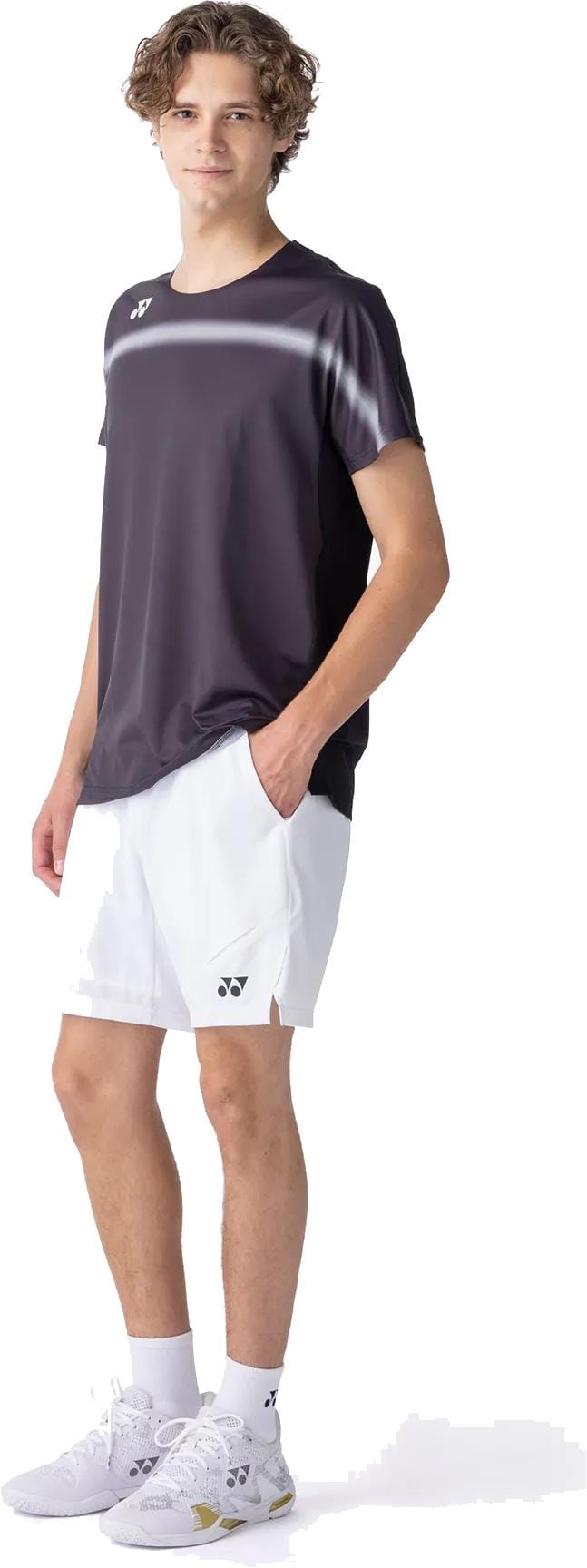 A person stands with one hand in their pocket, wearing a black Yonex 10711YX Crew Neck Mens Tennis T-Shirt, white shorts, socks, and athletic shoes, against a plain white background.