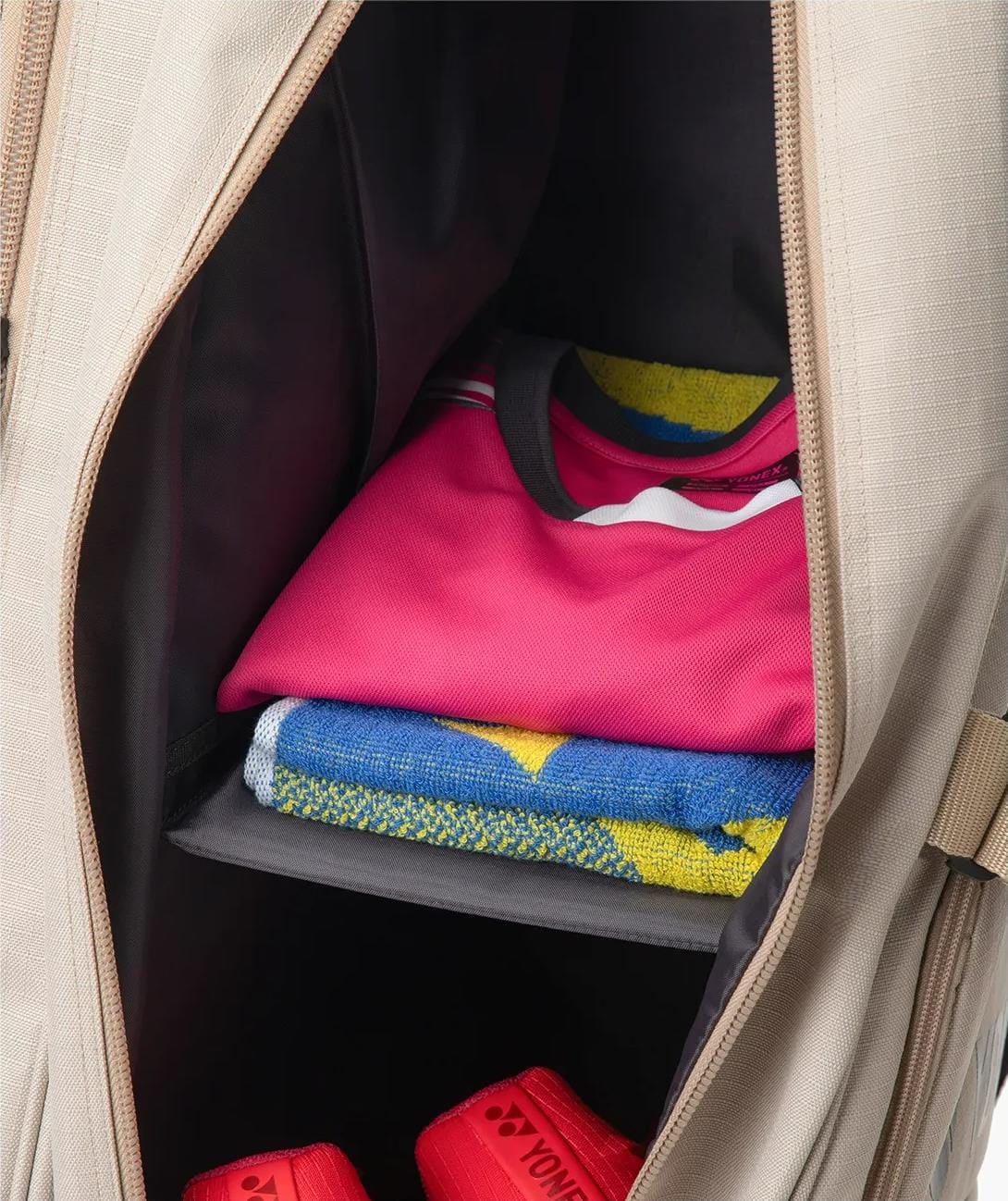 A spacious Yonex BA72626EX Gearlogic 6 Racket Tennis Bag in beige, made from recycled polyester, displays neatly folded clothes, a pink t-shirt, a colorful towel, and red Yonex shoes on its bottom shelf.