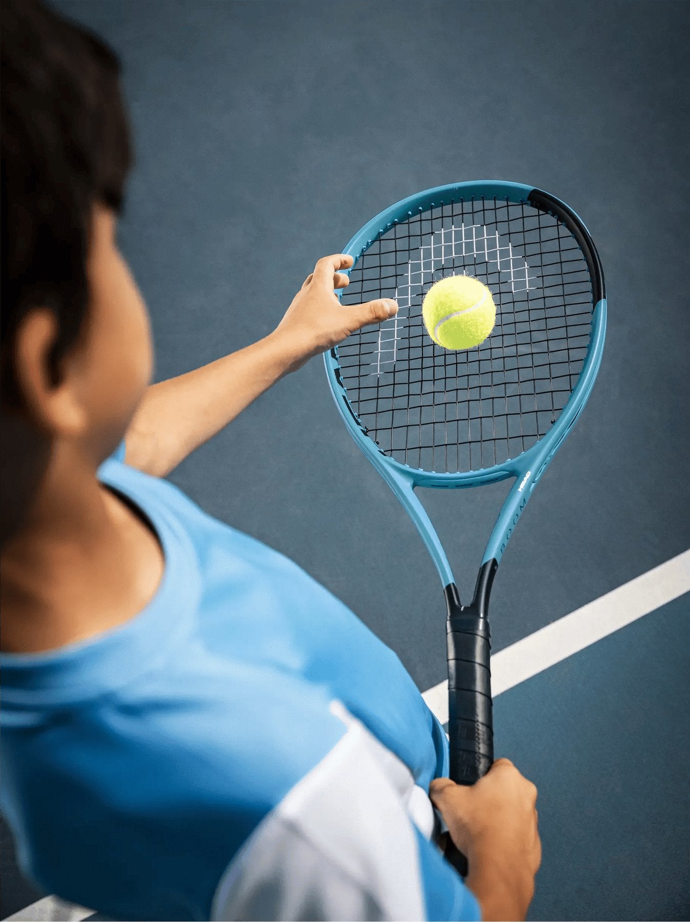 Wearing a blue shirt, a person prepares to serve on a blue tennis court, tossing a yellow ball above their HEAD Boom Jr 2026 Junior Tennis Racket by HEAD.