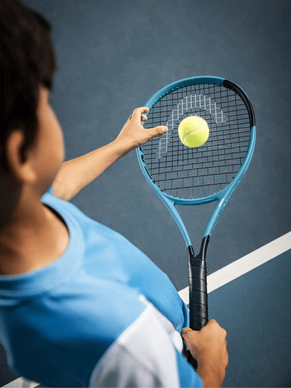 Wearing a blue shirt, a person prepares to serve on a blue tennis court, tossing a yellow ball above their HEAD Boom Jr 2026 Junior Tennis Racket by HEAD.