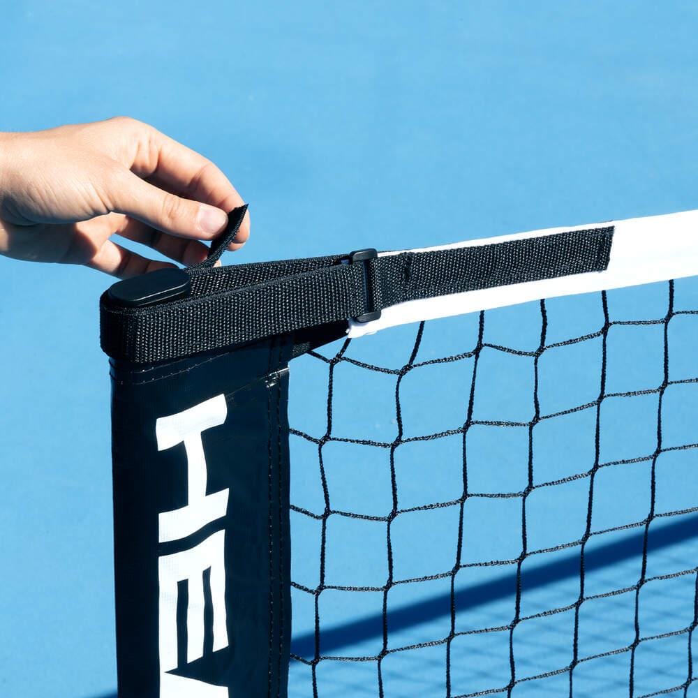 A hand adjusts the black strap and buckle at the top of a HEAD Portable Pickleball Net on a blue tennis court, with the HEAD logo partially visible on the net post cover.