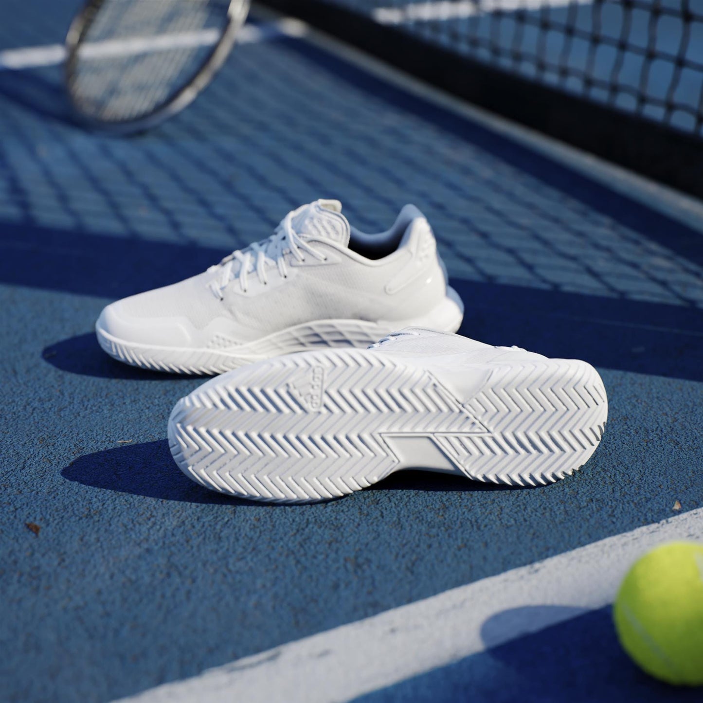A pair of adidas ADIDAS Defiant Speed 2 Womens Tennis Shoes in white on a blue court near a yellow ball, with a racket and net behind. These lightweight shoes deliver style and high performance for every match.