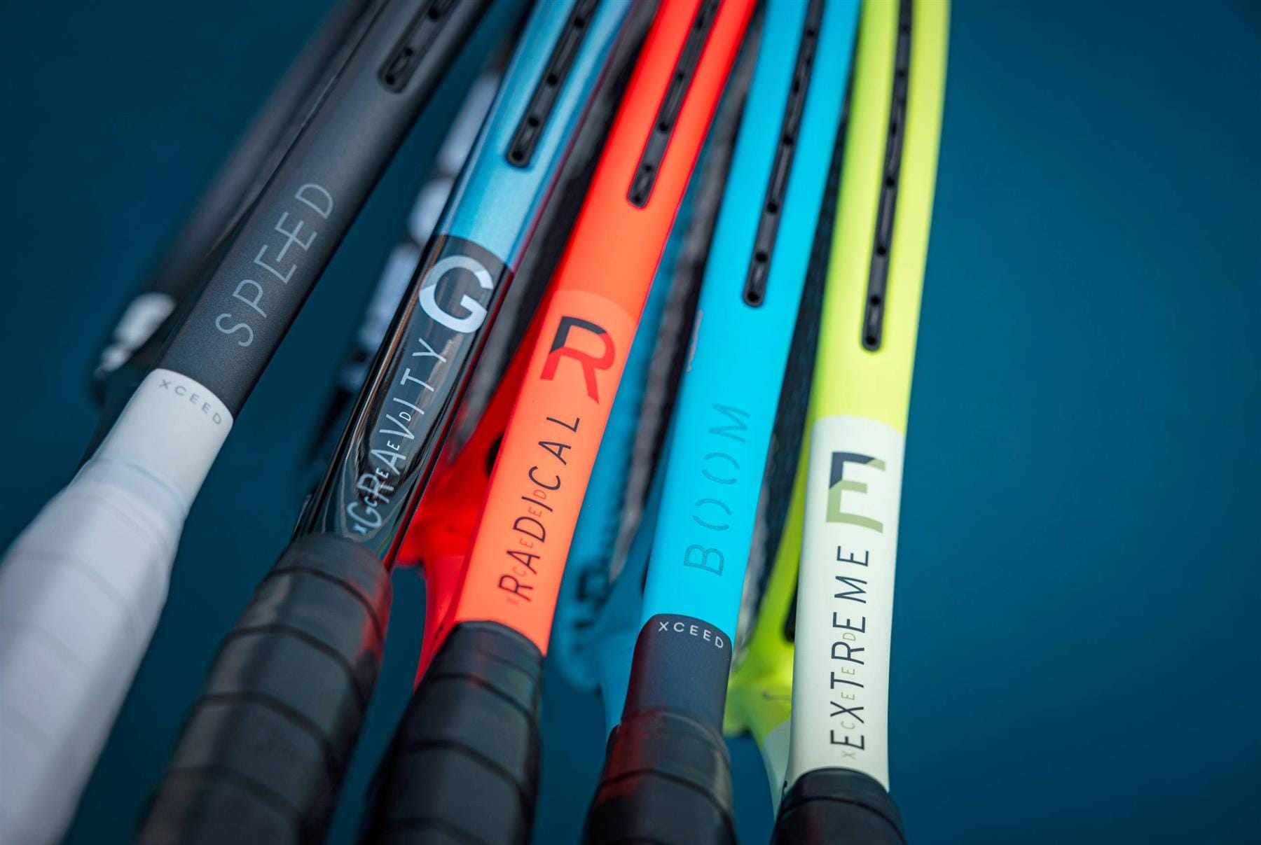 Close-up of the handles and lower frames of five HEAD graphite tennis rackets, including the HEAD IG Boom XCEED Tennis Racket, labeled SPEED, GRAVITY, RADICAL, BOOM, and EXTREME side by side on a blue surface.