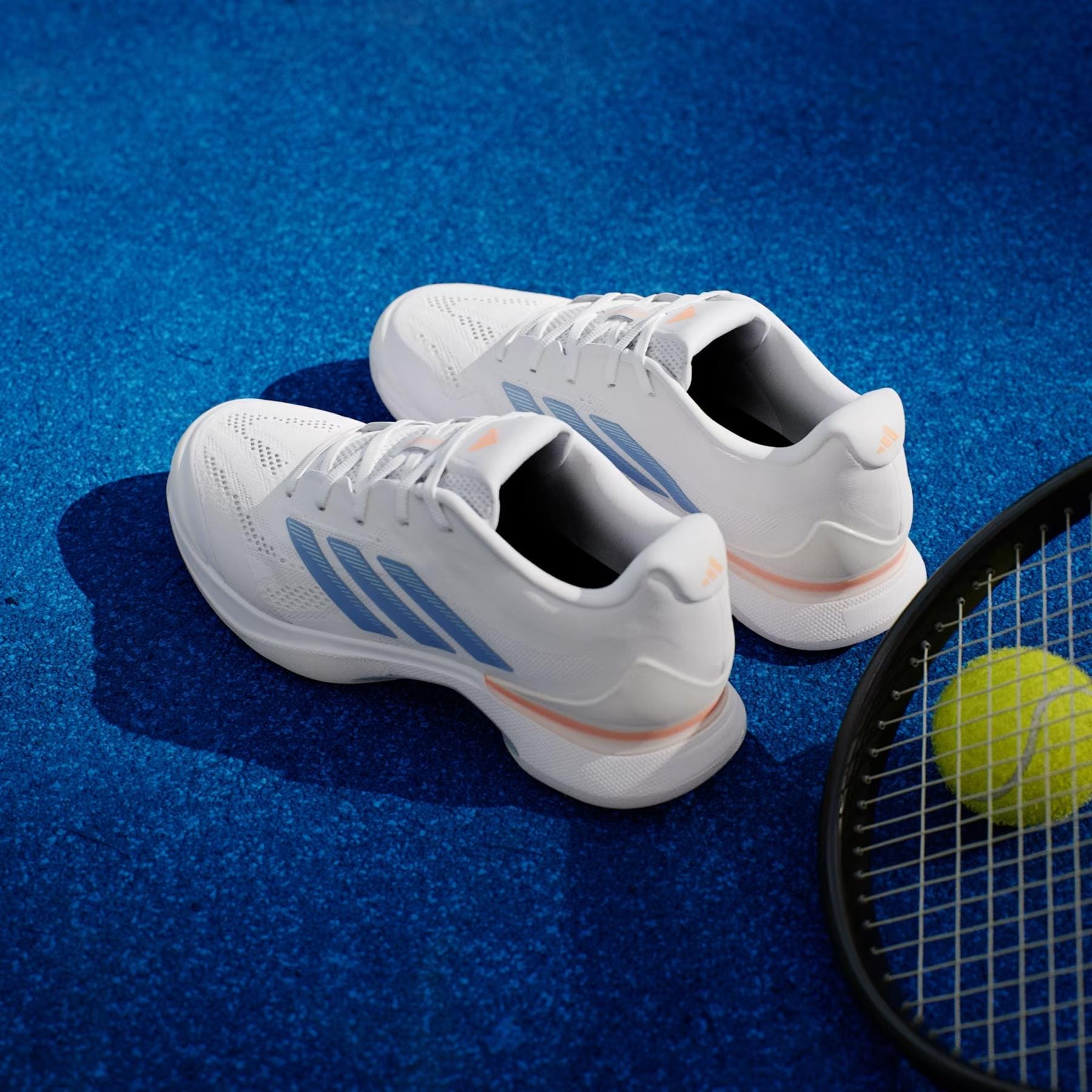 A pair of adidas Avacourt 3 Womens Tennis Shoes in Core White, Halo Blue, and Beam Orange are displayed on a blue tennis court beside a yellow tennis ball and the edge of a racket.