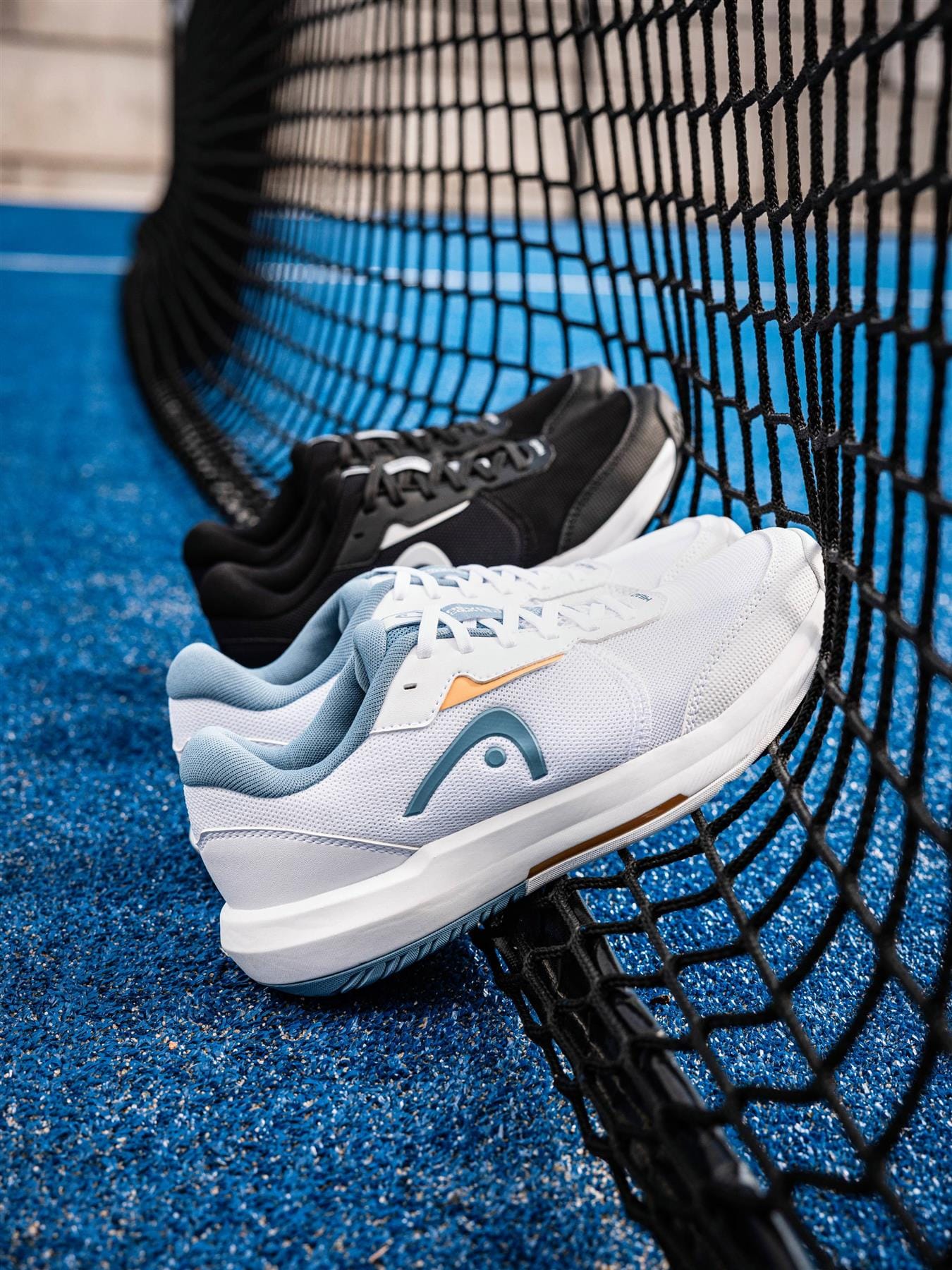 Two pairs of HEAD Revolt Court 5.0 Womens Tennis Shoes—one black, one white—rest against a black net on a bright blue court, showcasing the stylish performance of these HEAD tennis shoes in action.
