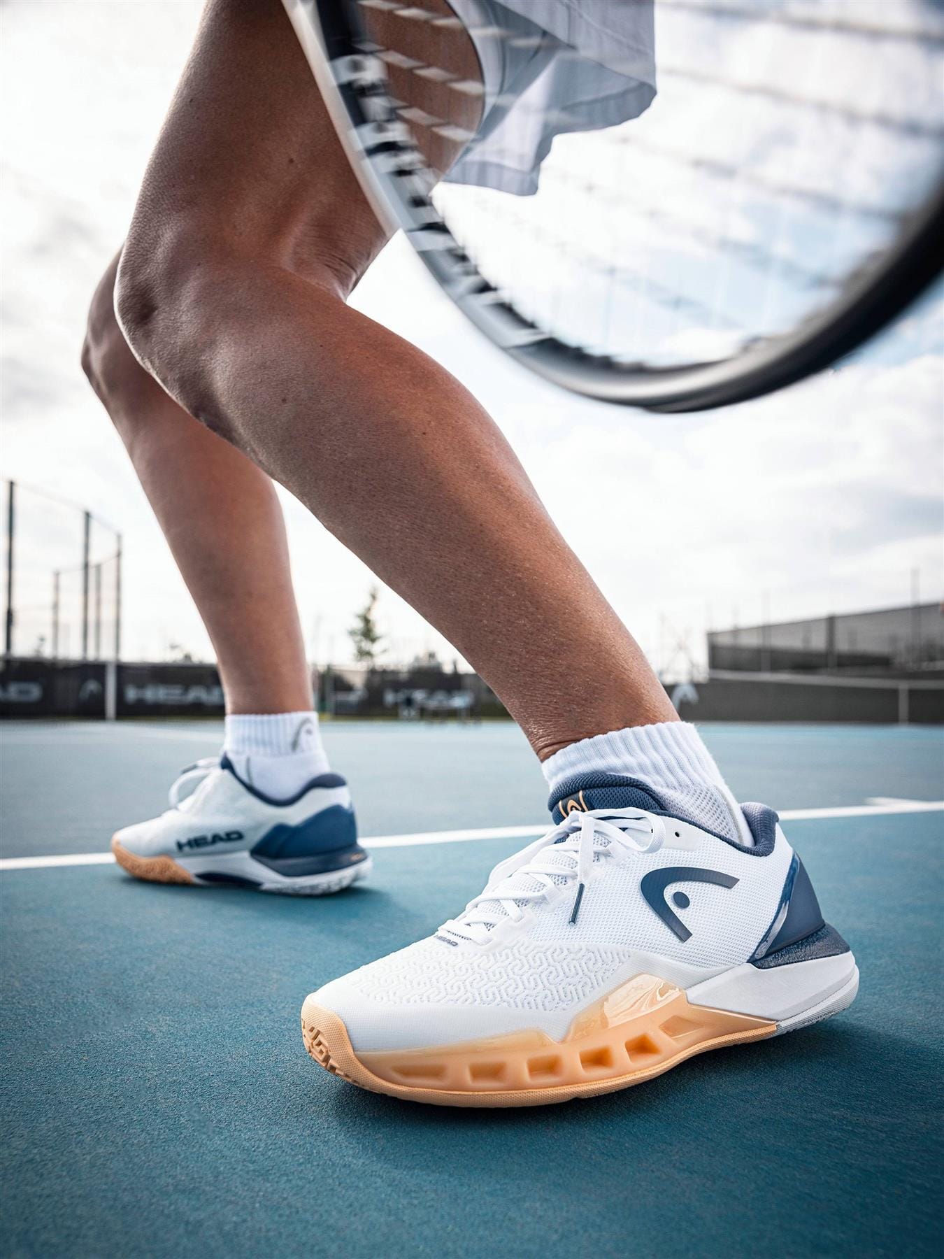 Close-up of a tennis player’s legs in an athletic stance on court, wearing HEAD Revolt Pro 5.0 Womens Tennis Shoes in White/Apricot by HEAD, with a blurred racket in the foreground.