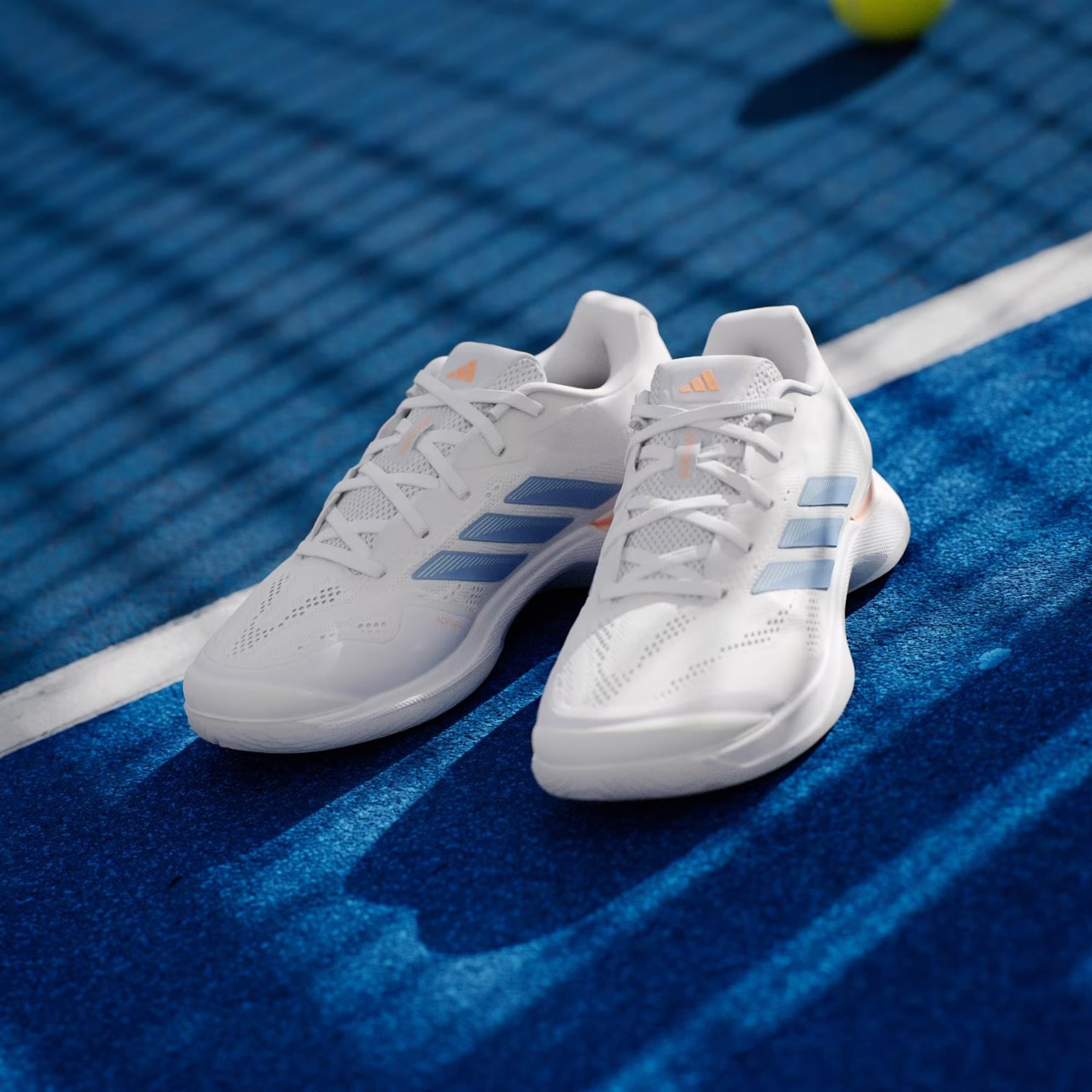 A pair of adidas Avacourt 3 Womens Tennis Shoes in Core White, Halo Blue, and Beam Orange rests on a blue tennis court with a blurred tennis ball and net in the background.
