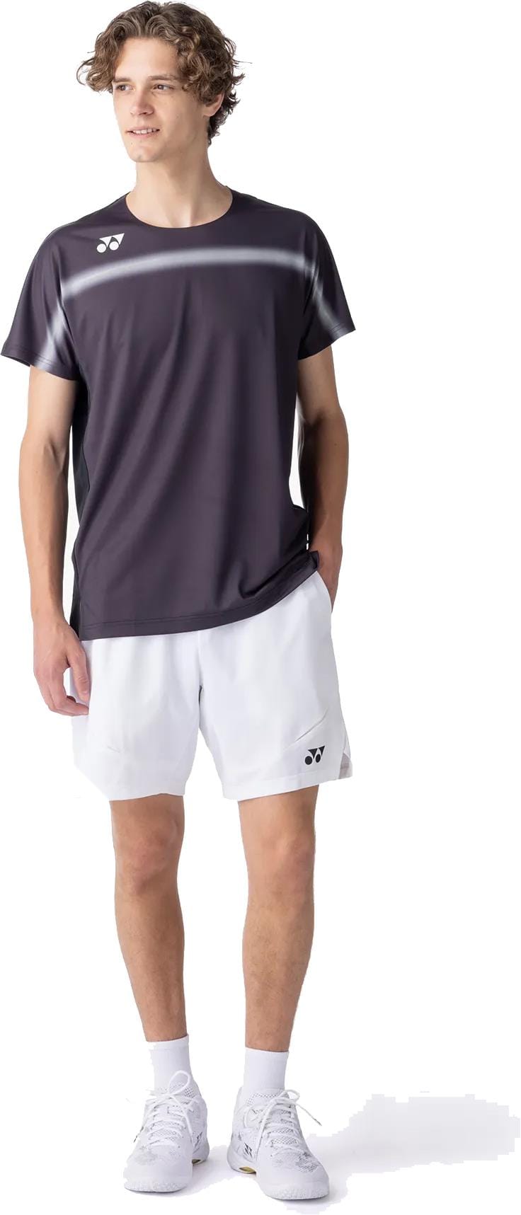 A young man with short curly hair wears a Yonex 10711YX Crew Neck Mens Tennis T-Shirt in black, paired with white athletic shorts, socks, and sneakers. He stands with one hand in his pocket, looking off to the side.
