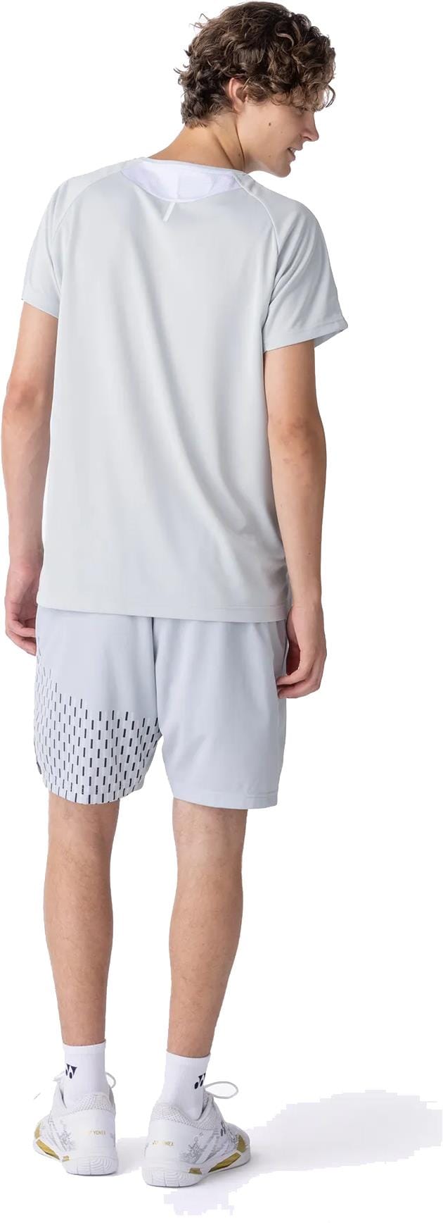 A person with curly hair is seen from behind wearing a Yonex 10713EX Crew Neck Mens Tennis T-Shirt in Silver Grey, matching dotted athletic shorts, white socks, and white sneakers.