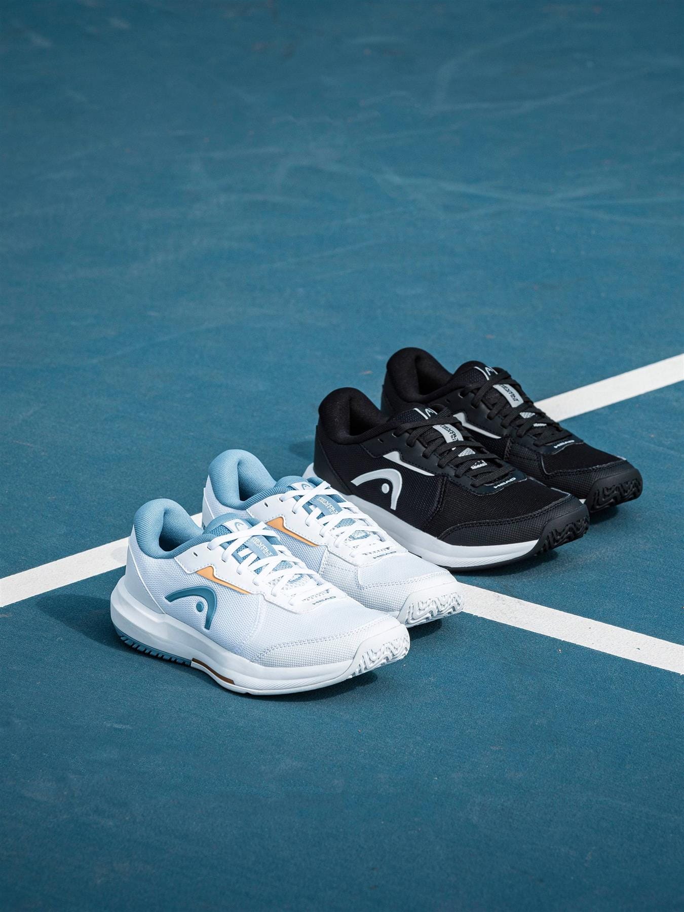 Three pairs of HEAD Revolt Court 5.0 womens tennis shoes sit on a blue court—one in black and white, one in white with blue accents, and one in white with orange accents—all near the white court line.