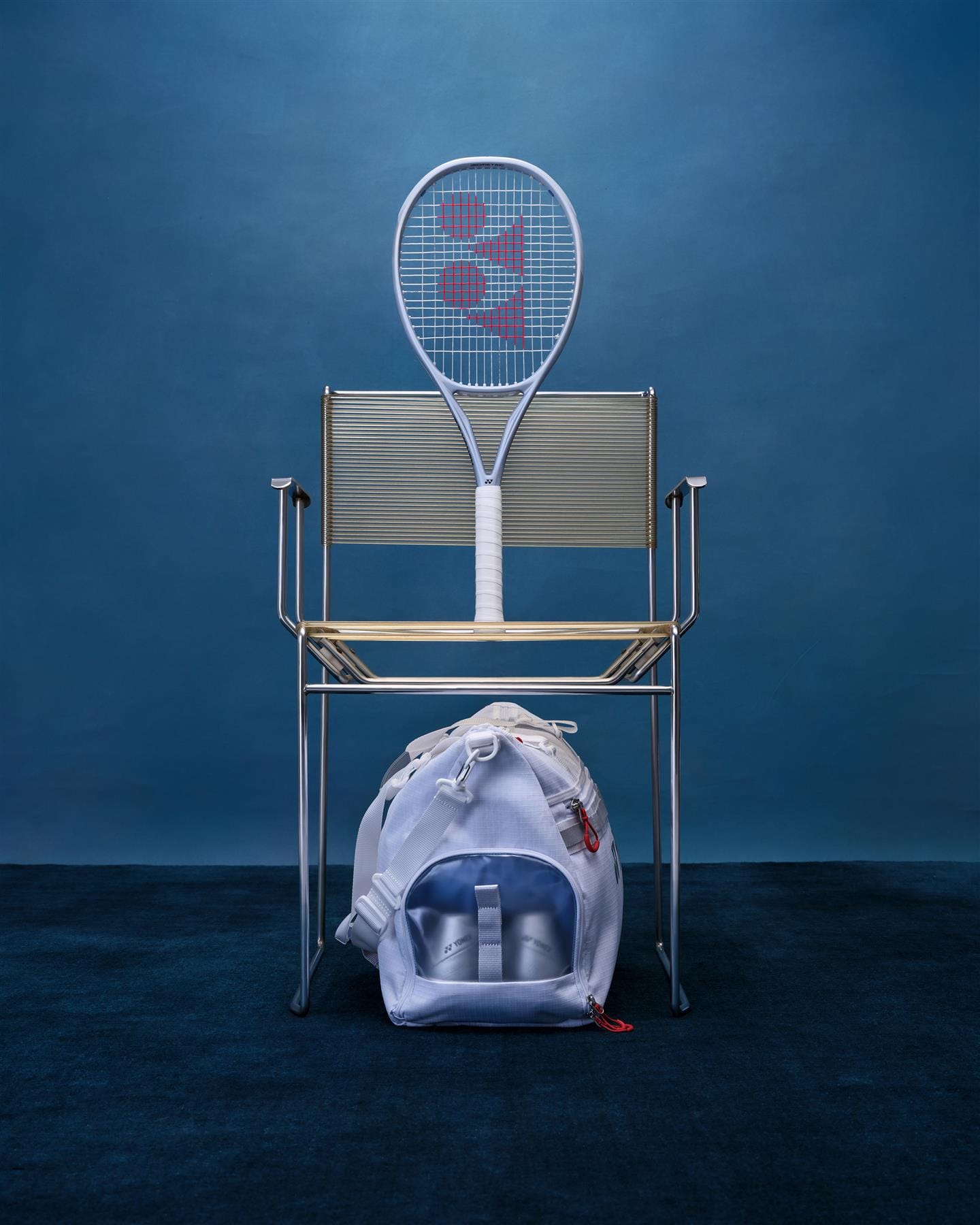 A Yonex Muse 100 Tennis Racket by Yonex stands upright on a sleek metal chair next to a white sports bag with bottles, all set against a blue backdrop—creating an inviting atmosphere for easy rallying.