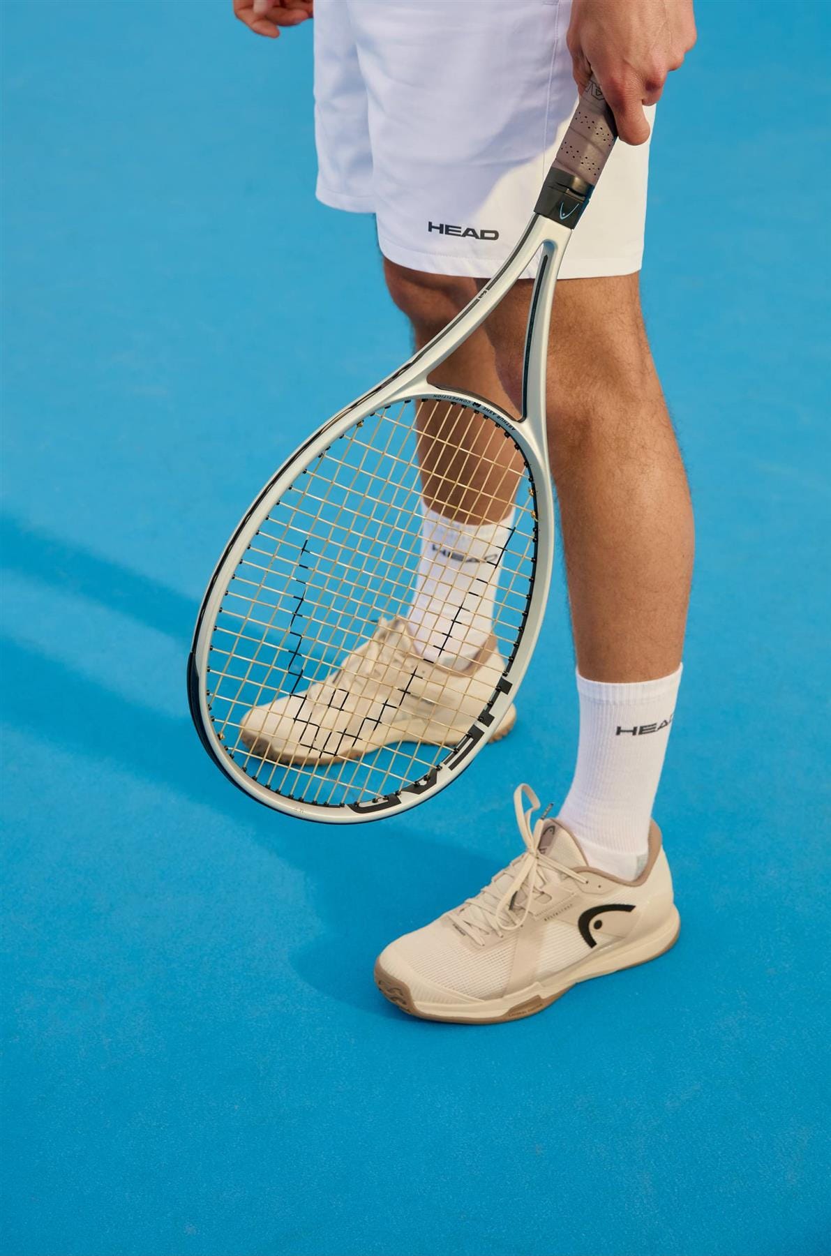 A tennis player’s lower body is visible on a bright blue court, wearing white shorts, socks, and HEAD Sprint Pro 4.0 Men's Tennis Shoes in Chalk White/Dark Grey while holding a racket.