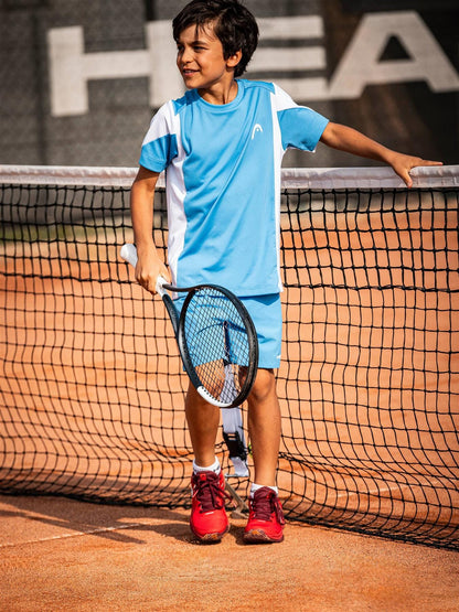 A young boy in blue sportswear stands by a tennis net on a clay court, smiling and holding a HEAD Speed Junior 25 2026 Tennis Racket, with the HEAD brand visible in the background.