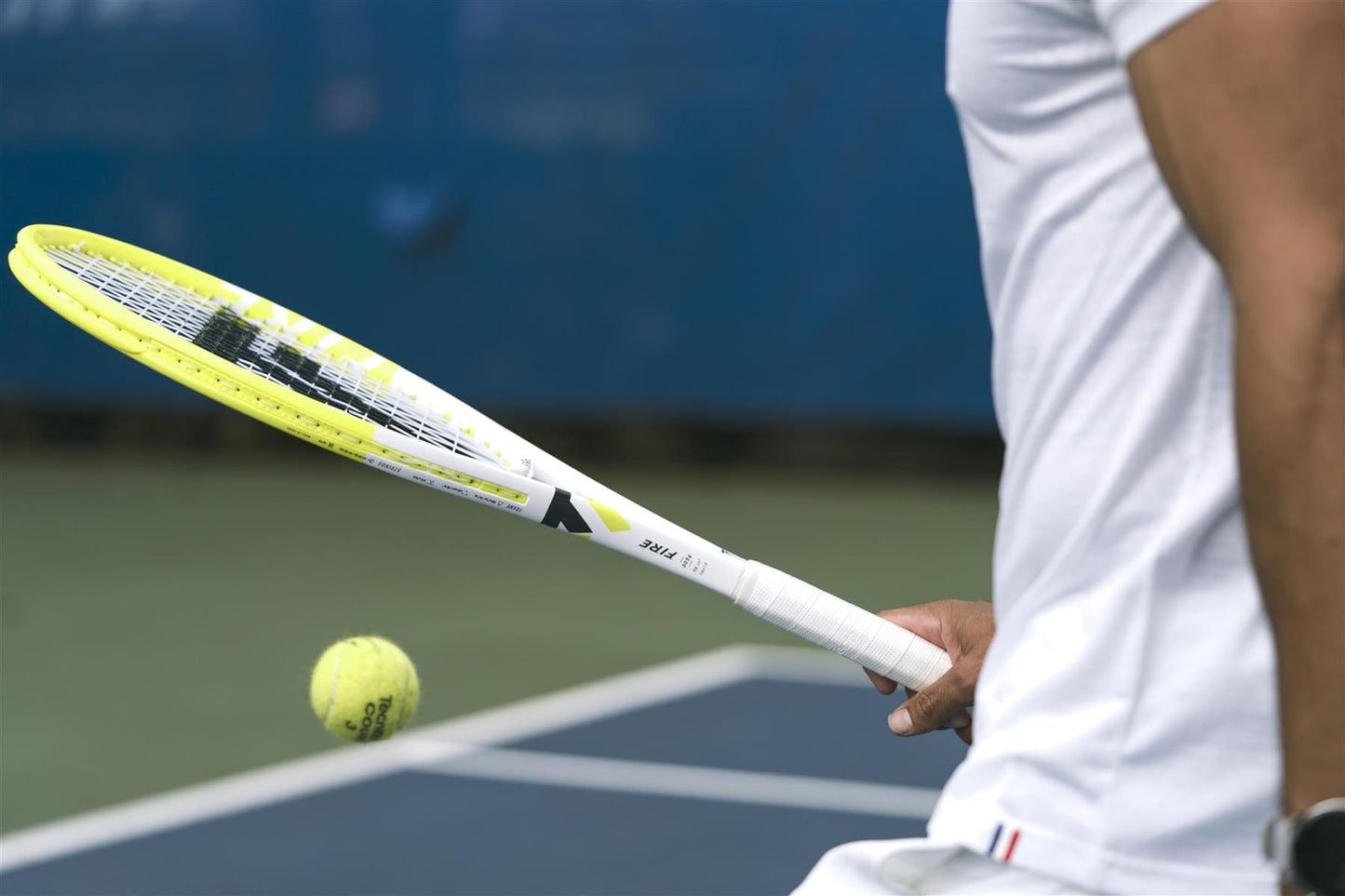Wearing a white shirt, a person prepares to hit a tennis ball on an outdoor court with a blue background, holding the Tecnifibre Fire 255 Tennis Racket by Tecnifibre featuring X-Damp technology.