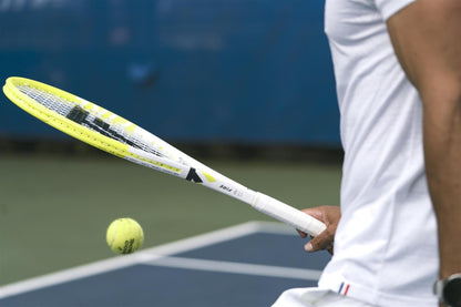 Wearing a white shirt, a person prepares to hit a tennis ball on an outdoor court with a blue background, holding the Tecnifibre Fire 255 Tennis Racket by Tecnifibre featuring X-Damp technology.