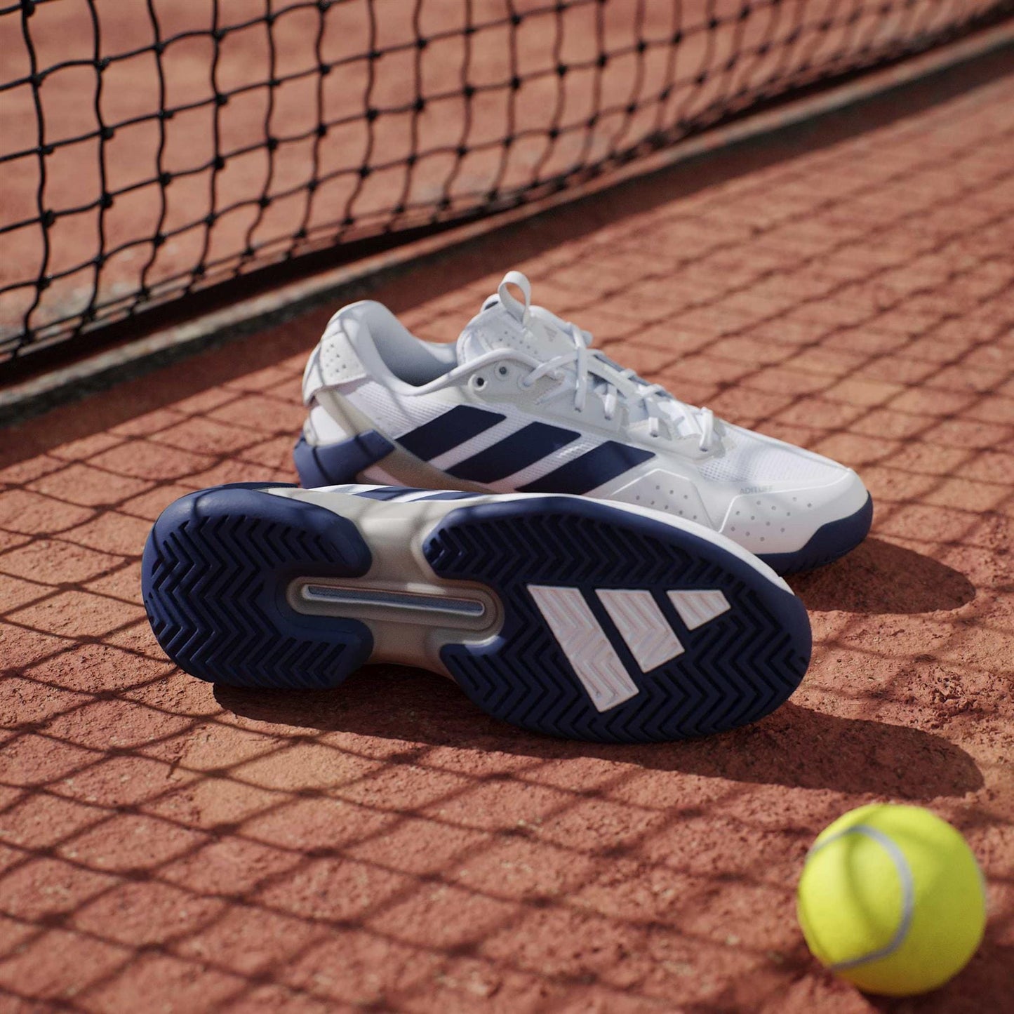A pair of lightweight adidas Adizero Ubersonic 5 Men's Tennis Shoes in White/Navy rest on a clay court beside a tennis ball, with a net visible in the background.