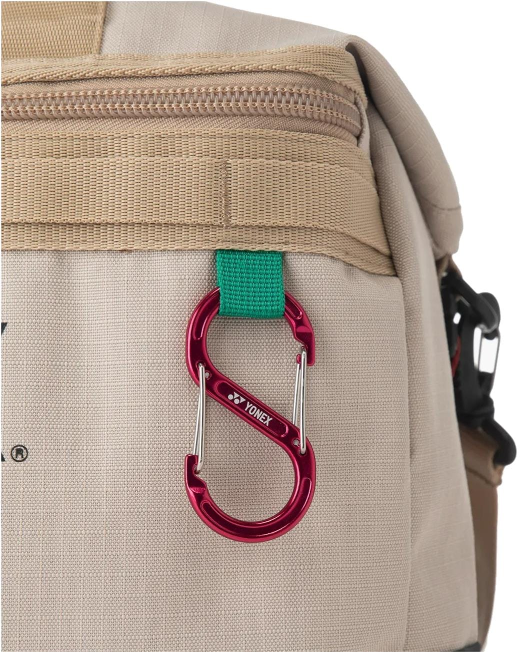 A red Yonex S-shaped carabiner is attached to a green fabric loop on the Yonex BA72611EX Gearlogic Tennis Boston Bag in beige, which features zippers and multiple compartments.