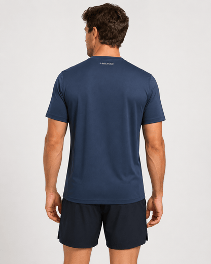 A man stands with his back to the camera, wearing a HEAD Club Tech 25 Men’s Tennis T-Shirt in navy and black shorts against a plain light background. The word “HEAD” appears near the neckline on the back of the shirt. This breathable tennis tee from HEAD is designed with moisture transfer microfiber for enhanced comfort during intense matches.