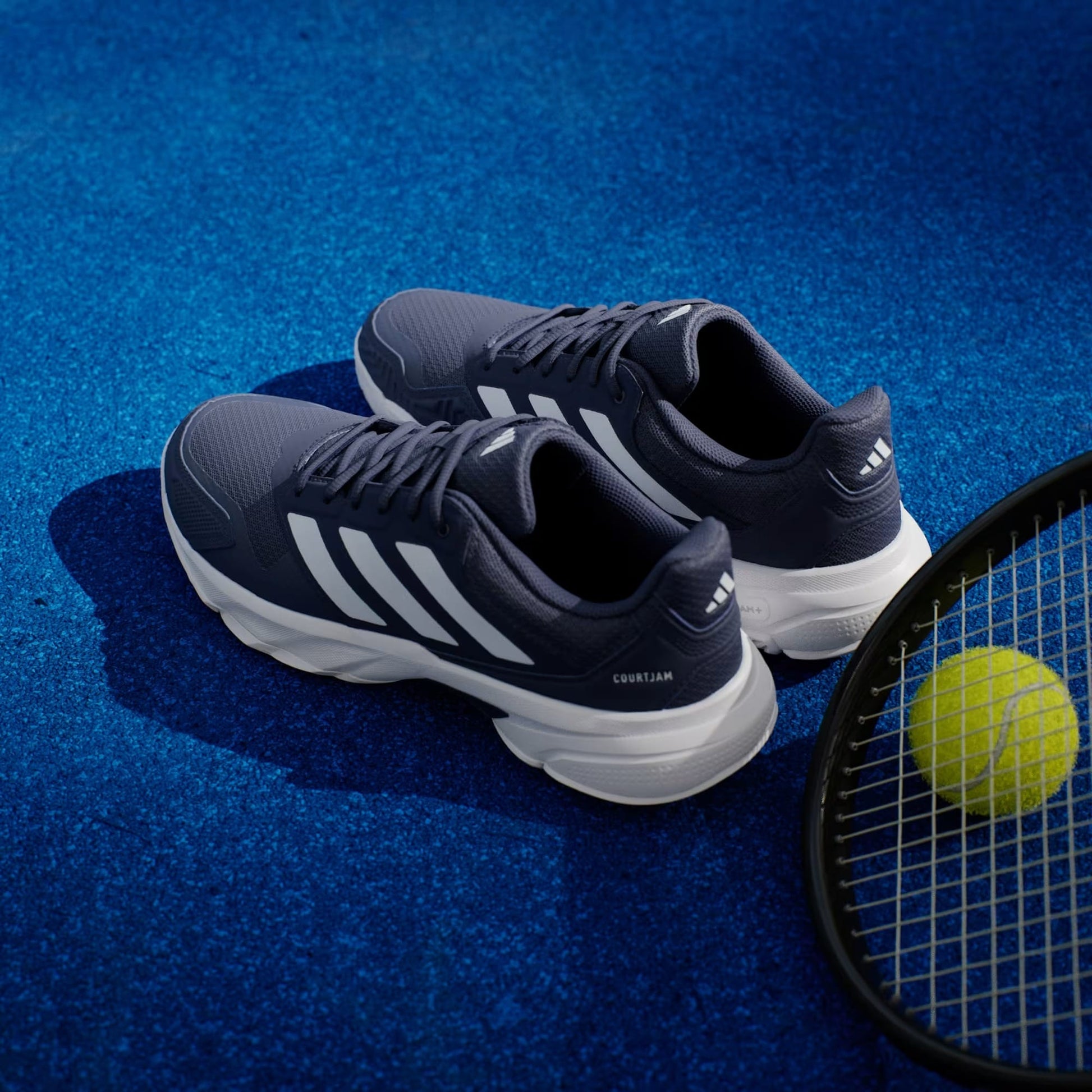 A pair of adidas CourtJam Control 3 Men’s Tennis Shoes in Dark Blue and Cloud White rests on a blue tennis court beside a tennis racket and a yellow ball, delivering the stability you need for every match.