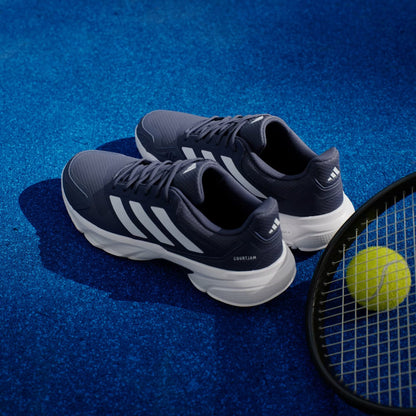 A pair of adidas CourtJam Control 3 Men’s Tennis Shoes in Dark Blue and Cloud White rests on a blue tennis court beside a tennis racket and a yellow ball, delivering the stability you need for every match.