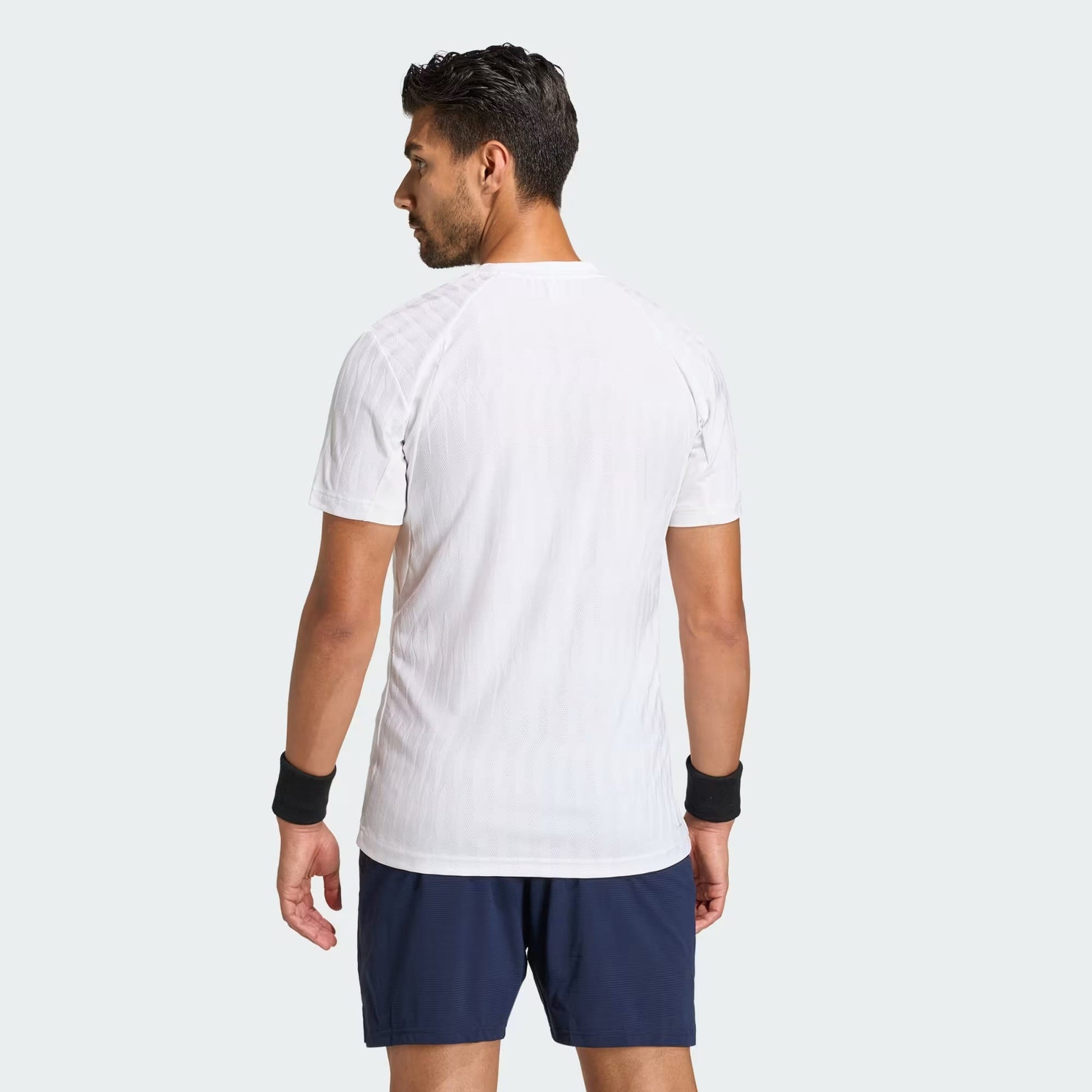 A man faces away from the camera, wearing an adidas Climacool+ Airchill Freelift Men's Tennis T-Shirt in white, navy shorts, and black wristbands, set against a plain light gray background.
