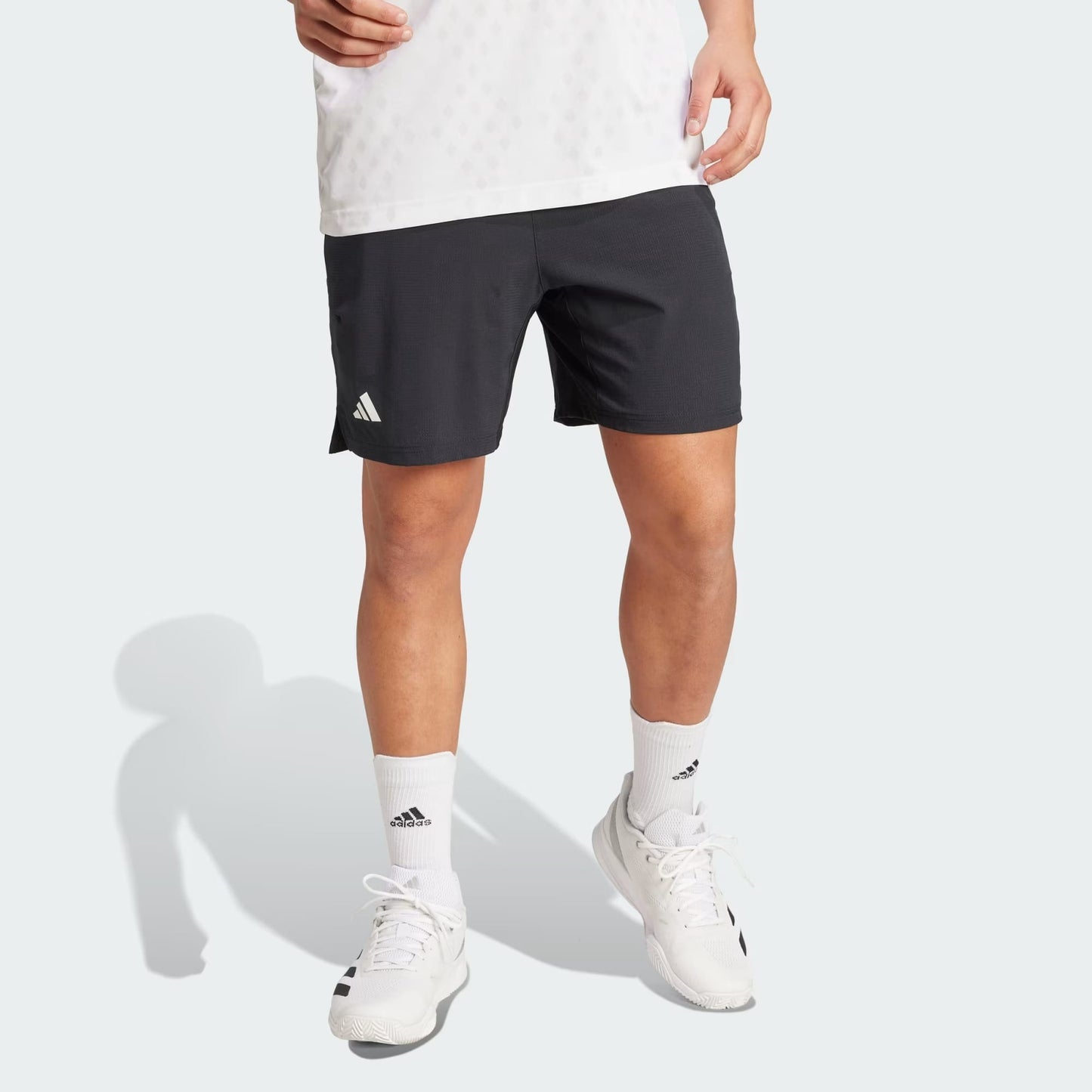 A person’s lower body is shown wearing adidas Climacool Ergo Mens Tennis Shorts in black, paired with a white shirt, white adidas socks, and white athletic shoes on a light background. The sweat-wicking shorts deliver comfort and style.