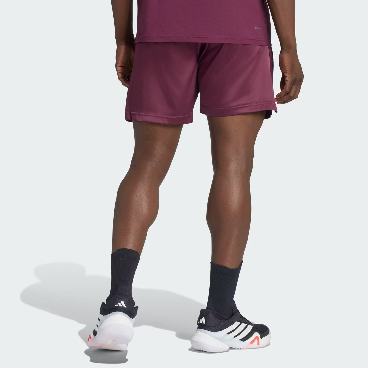 A person wearing adidas Climacool Ergo Pro Mens Tennis Shorts in maroon, a maroon shirt, black adidas socks, and black adidas sneakers with white soles stands with their back to the camera against a plain background.