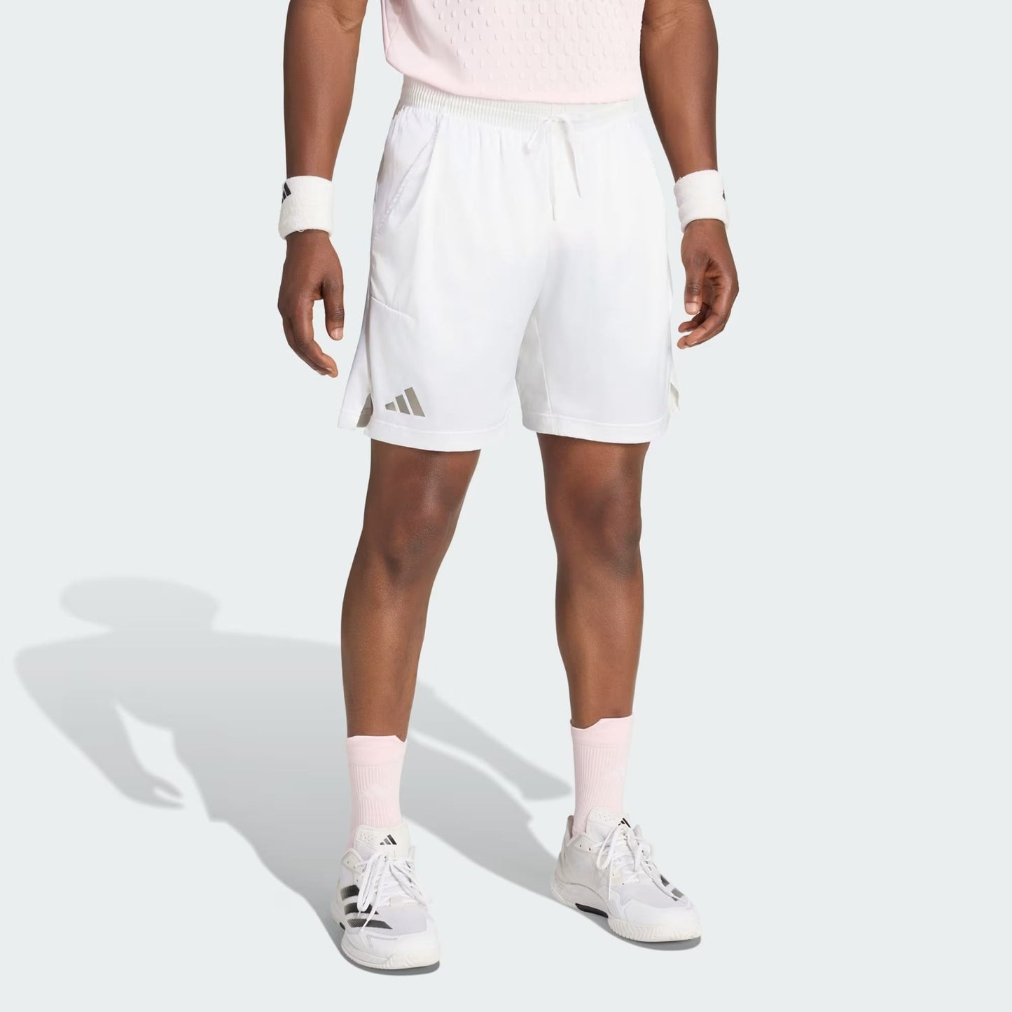 A person stands against a plain light background wearing adidas Climacool Ergo Pro Mens Tennis Shorts in white, paired with a light pink shirt, white wristbands, pink socks, and white sneakers. Their head is not visible in the image.