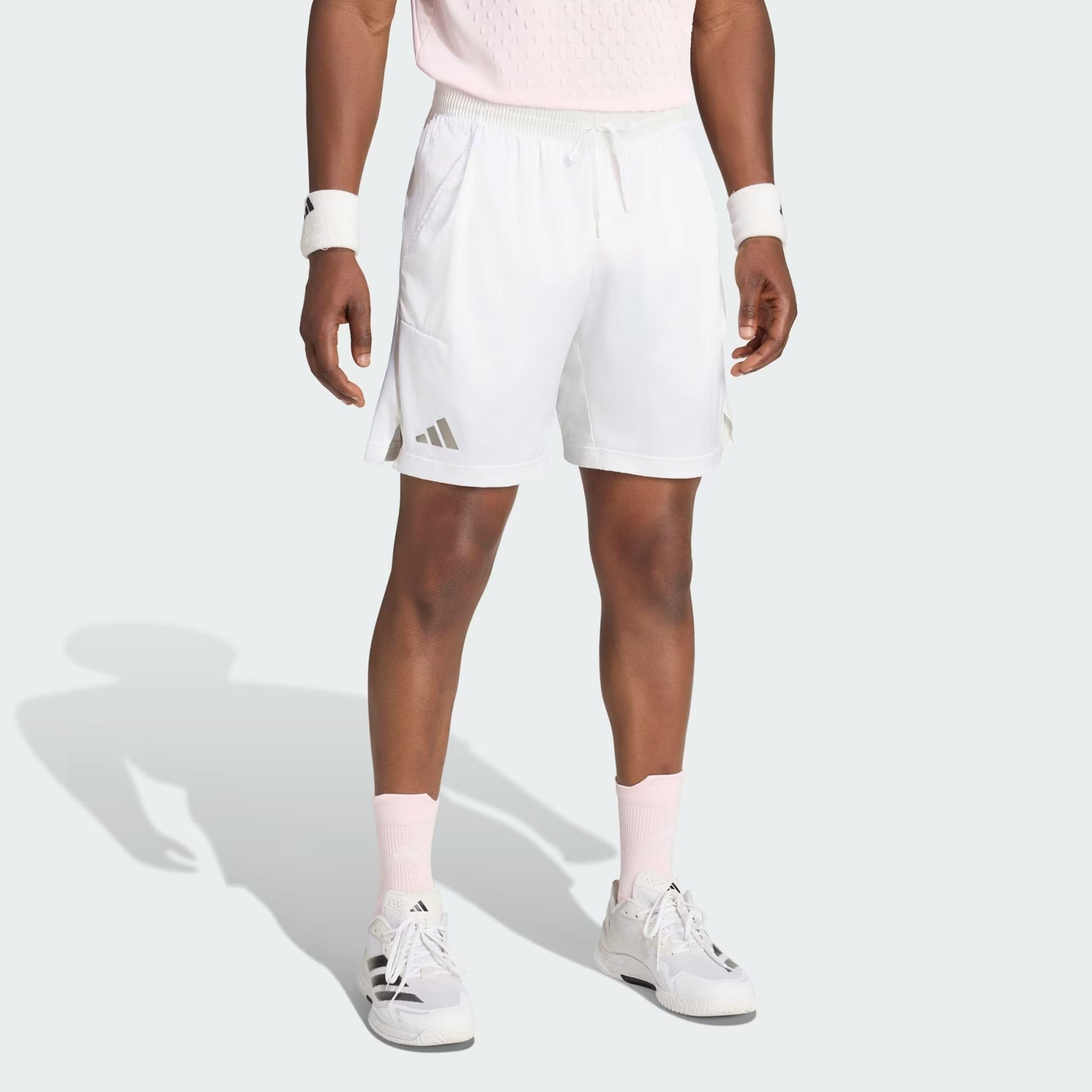 A person stands against a plain light background wearing adidas Climacool Ergo Pro Mens Tennis Shorts in white, paired with a light pink shirt, white wristbands, pink socks, and white sneakers. Their head is not visible in the image.