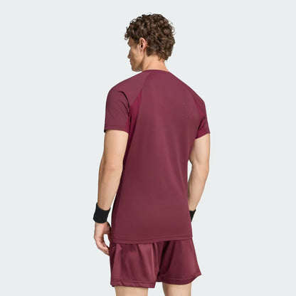 A man with curly hair stands facing away from the camera against a plain light background, wearing the adidas Climacool+ Freelift Pro Men’s Tennis T-Shirt in maroon along with matching shorts.