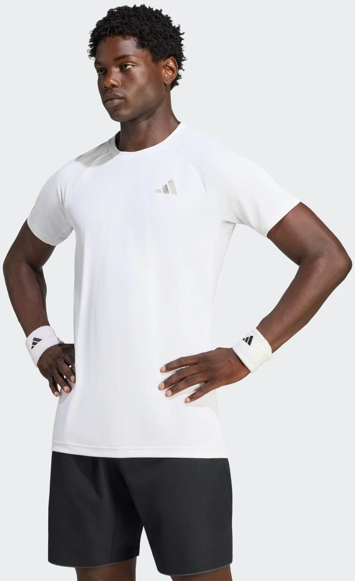 A man wearing the adidas Climacool+ Freelift Pro Men’s Tennis T-Shirt in white, paired with black shorts and white adidas wristbands, stands with his hands on his hips, looking to the side against a plain background.