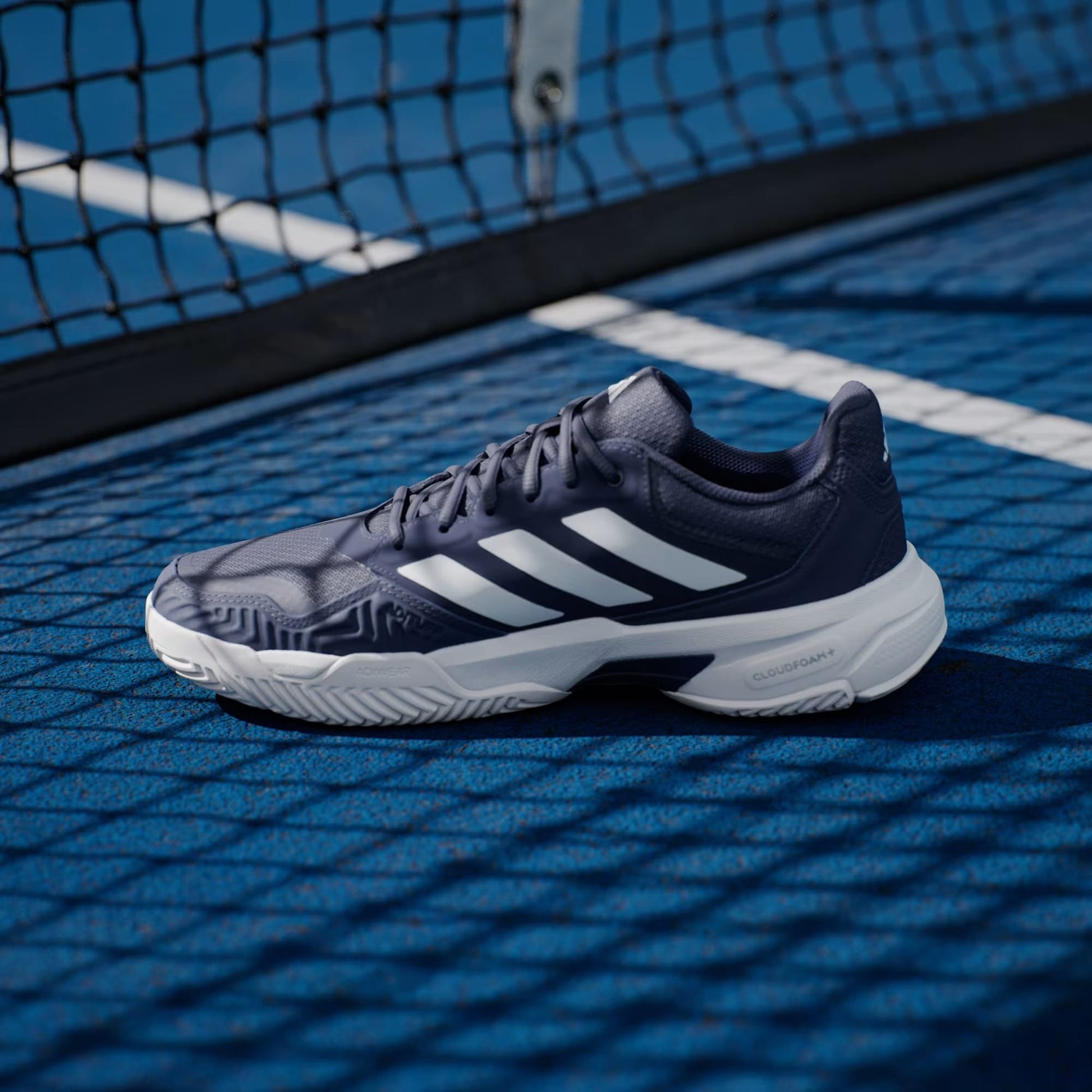 An adidas CourtJam Control 3 Mens Tennis Shoe in Dark Blue and Cloud White, recognized for its exceptional stability, featuring white stripes and a white sole, is placed on a blue tennis court near the net with visible white court lines.