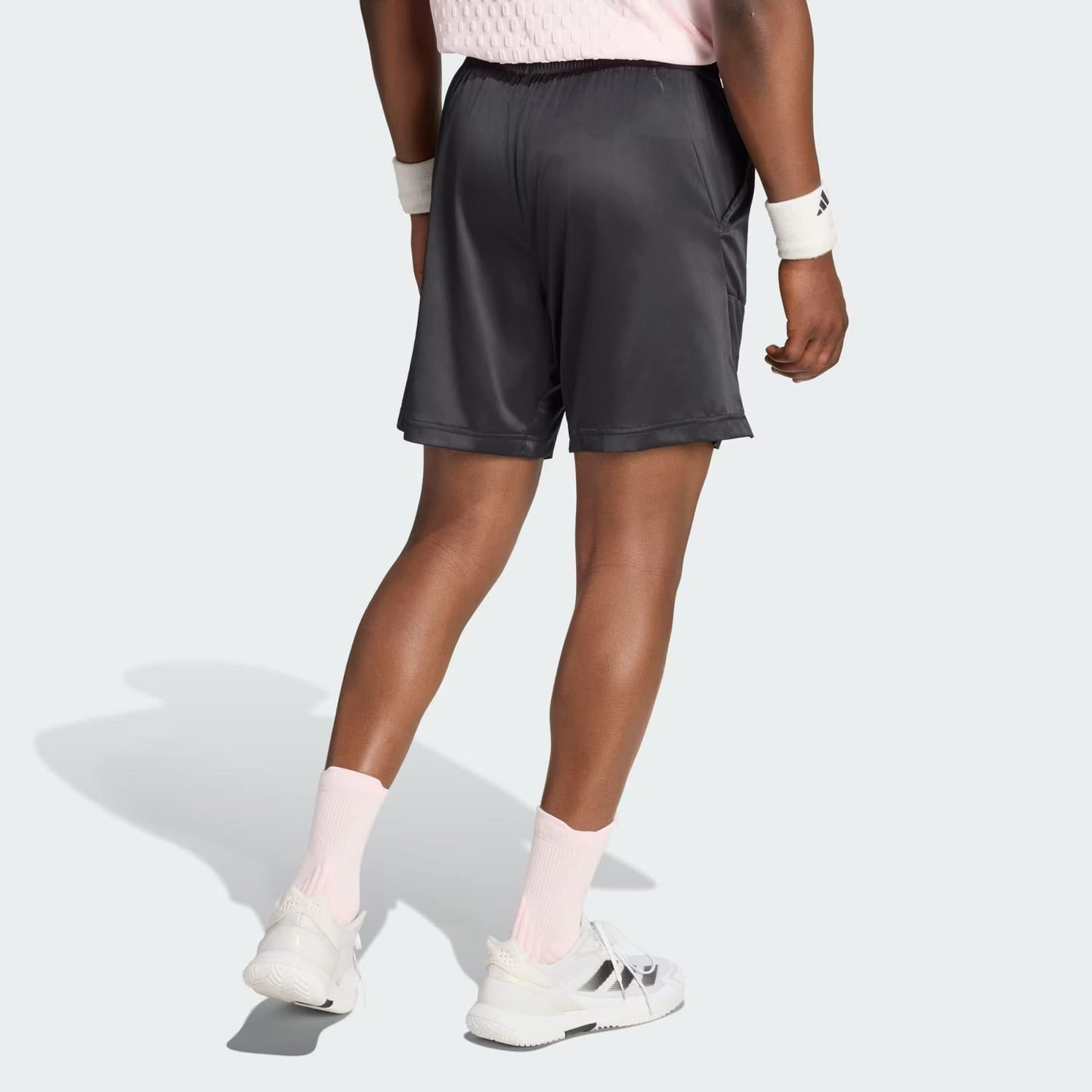 A person wearing a light pink shirt, adidas Climacool Ergo Pro Mens Tennis Shorts in black, pink socks, white sneakers, and white wristbands is shown standing with their back facing the camera on a light gray background.