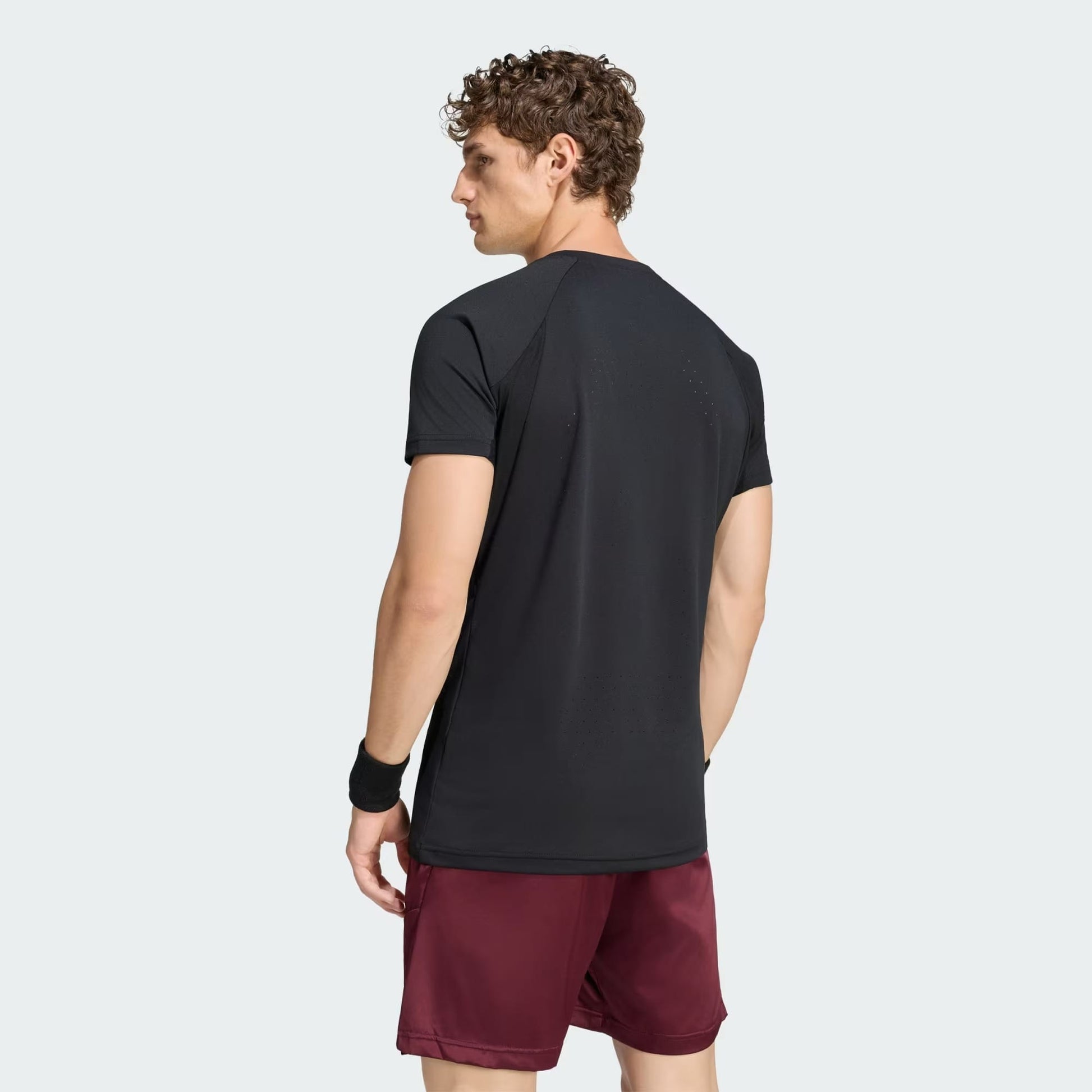 A man with curly hair is wearing an adidas Climacool+ Freelift Pro Men’s Tennis T-Shirt in black, burgundy shorts, and a black wristband. He stands facing away from the camera with a neutral expression.